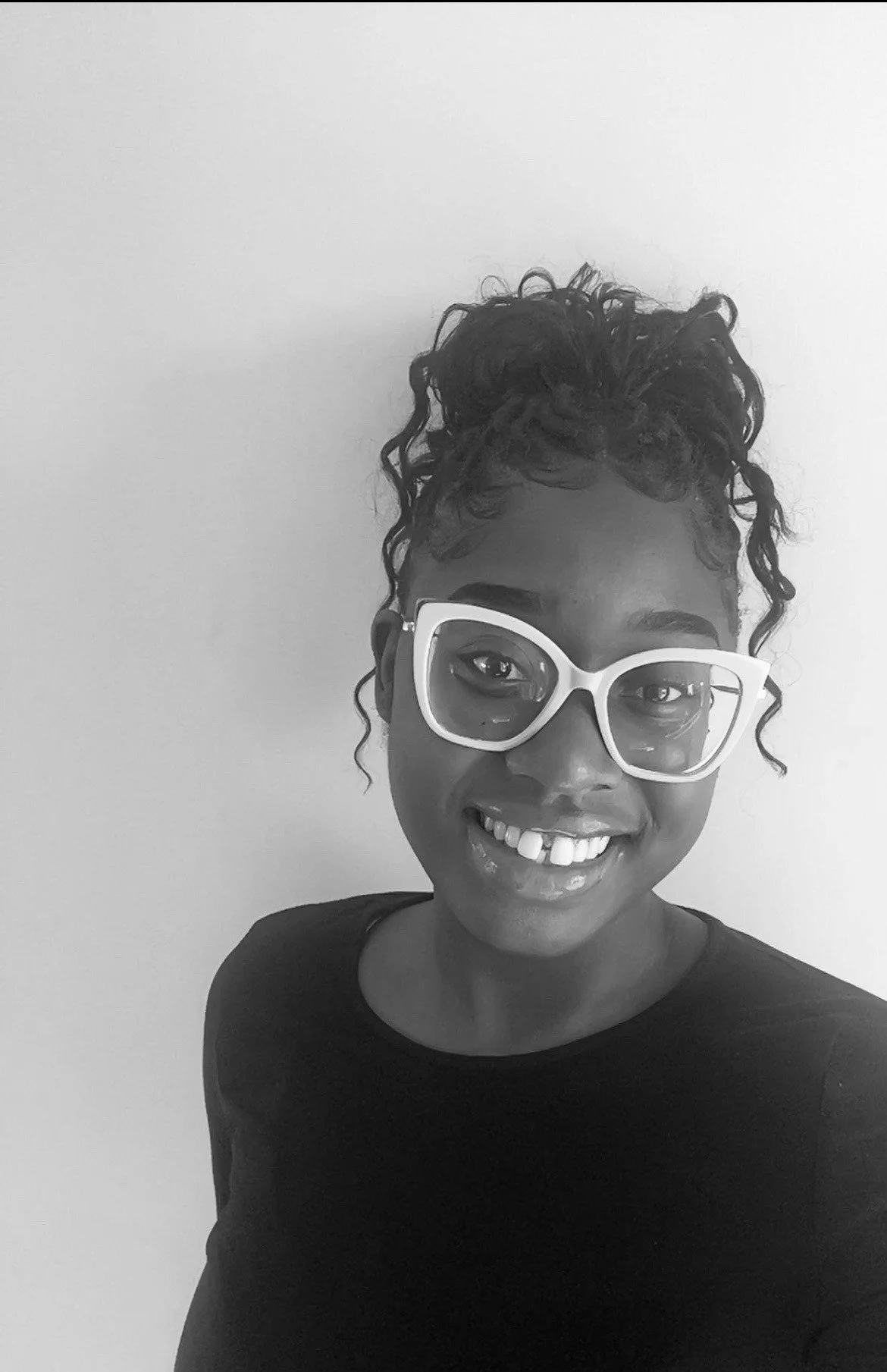 A woman with curly hair in a bun and wearing large glasses, smiling at the camera against a plain background.