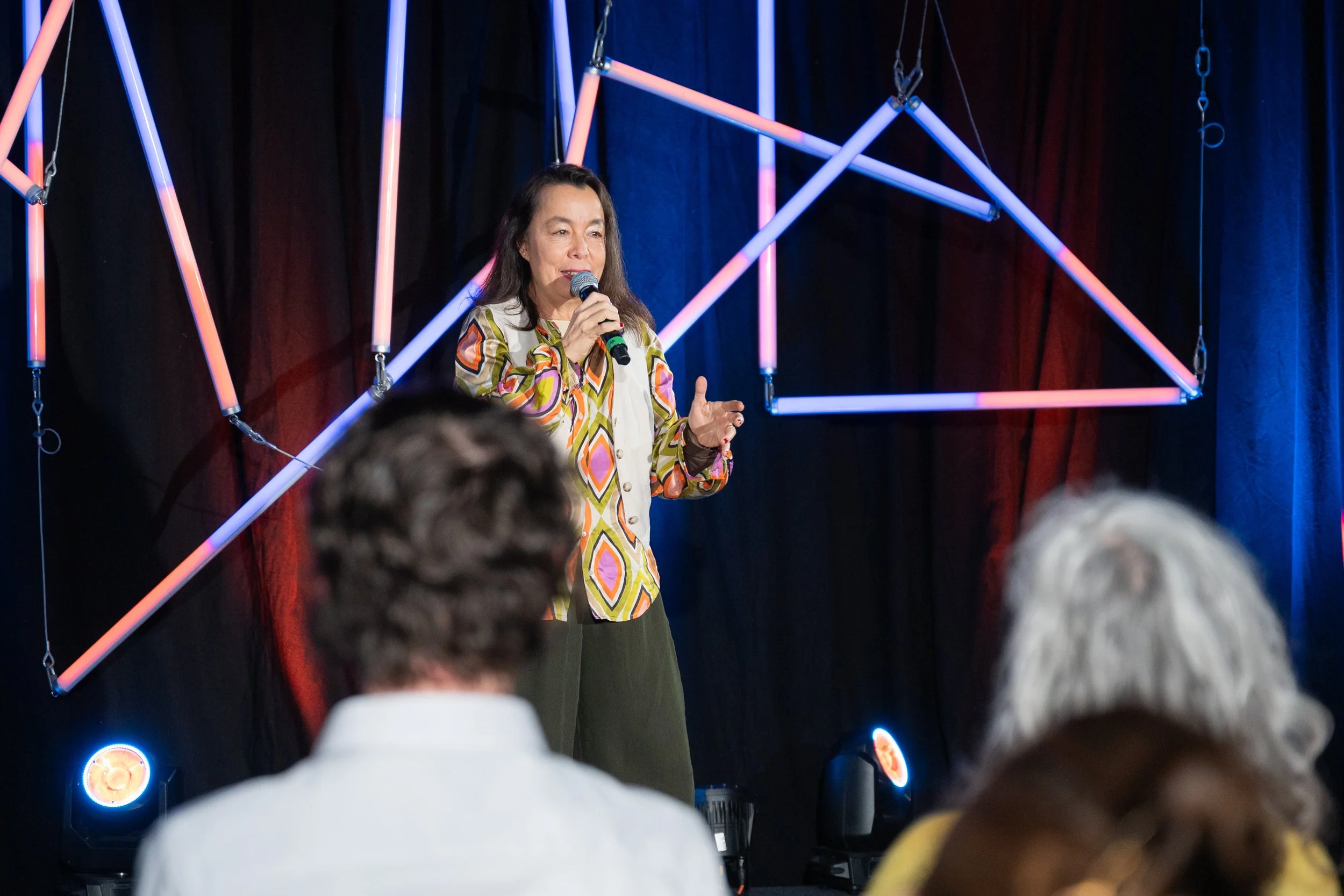 A woman speaking into a microphone on stage during a presentation or performance, with an audience in front of her, illuminated stage lights, and geometric shaped light fixtures hanging behind her.