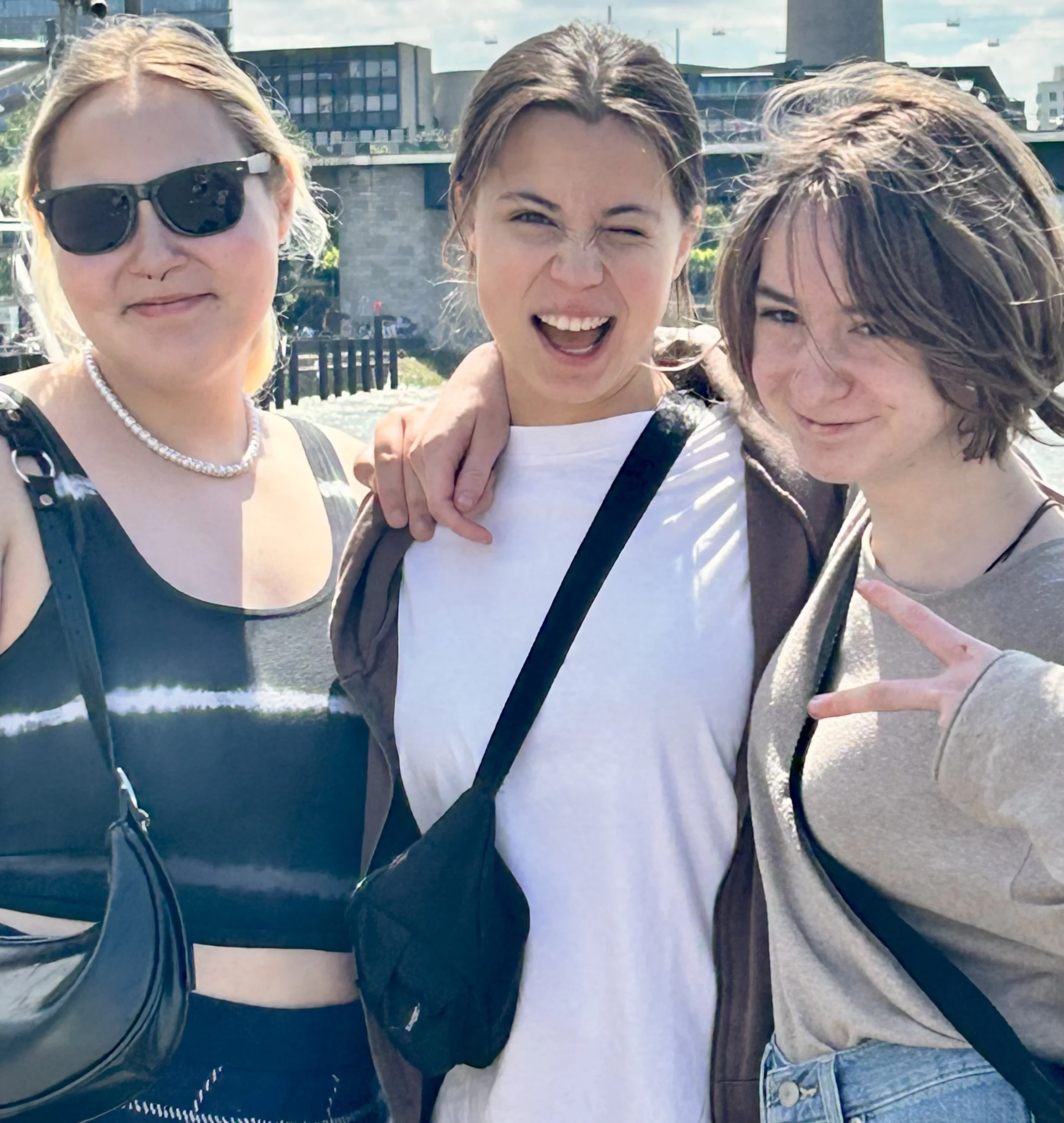 Three young women posing outdoors in a sunny urban environment, smiling and making playful expressions, with buildings and a cloudy sky in the background.
