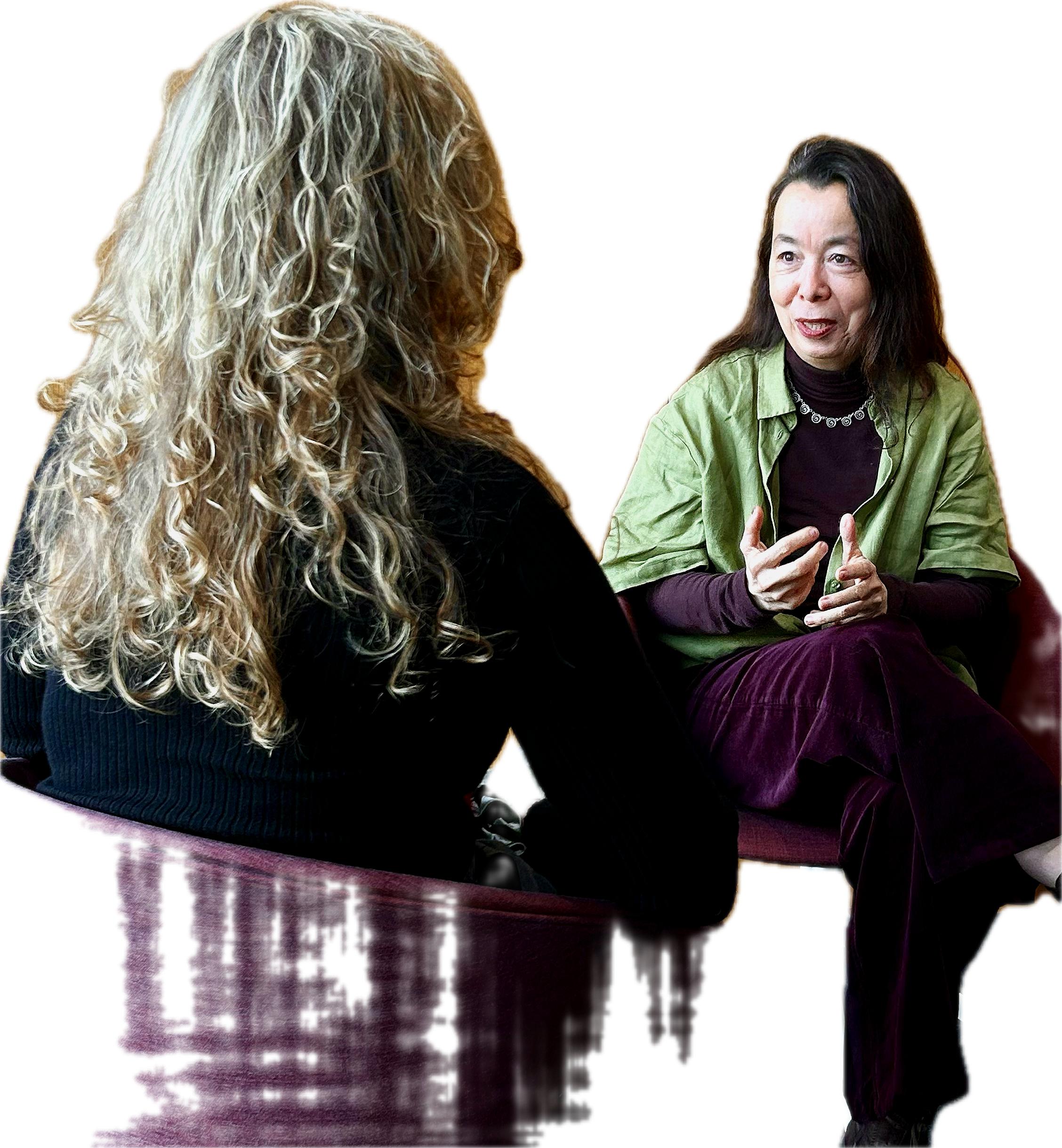Two women seated and talking, one with curly blonde hair wearing a black sweater, the other with black hair wearing a green shirt over a black top, against a bright yellow background.