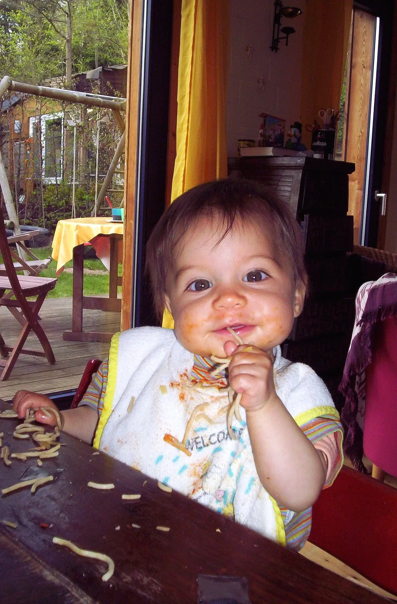 A smiling toddler with messy food on face and bib, sitting at a table eating noodles.