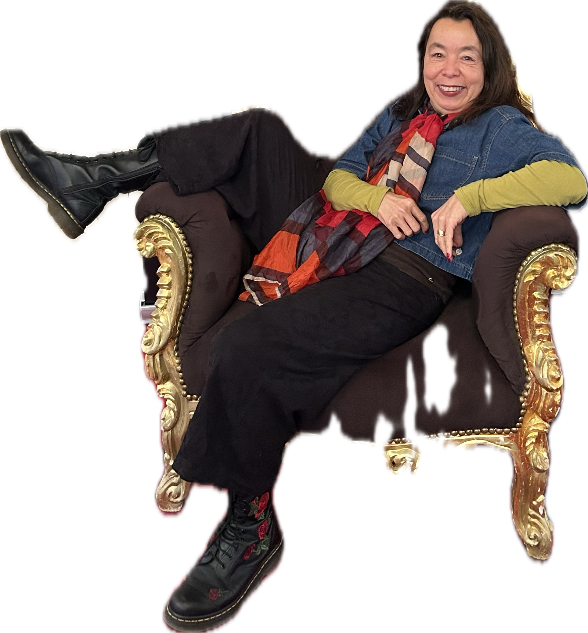 A woman with shoulder-length dark hair, wearing a denim jacket, a colorful scarf, black pants, and black boots, is lounging on a vintage, ornate, dark-colored sofa with gold accents, smiling and looking at the camera.