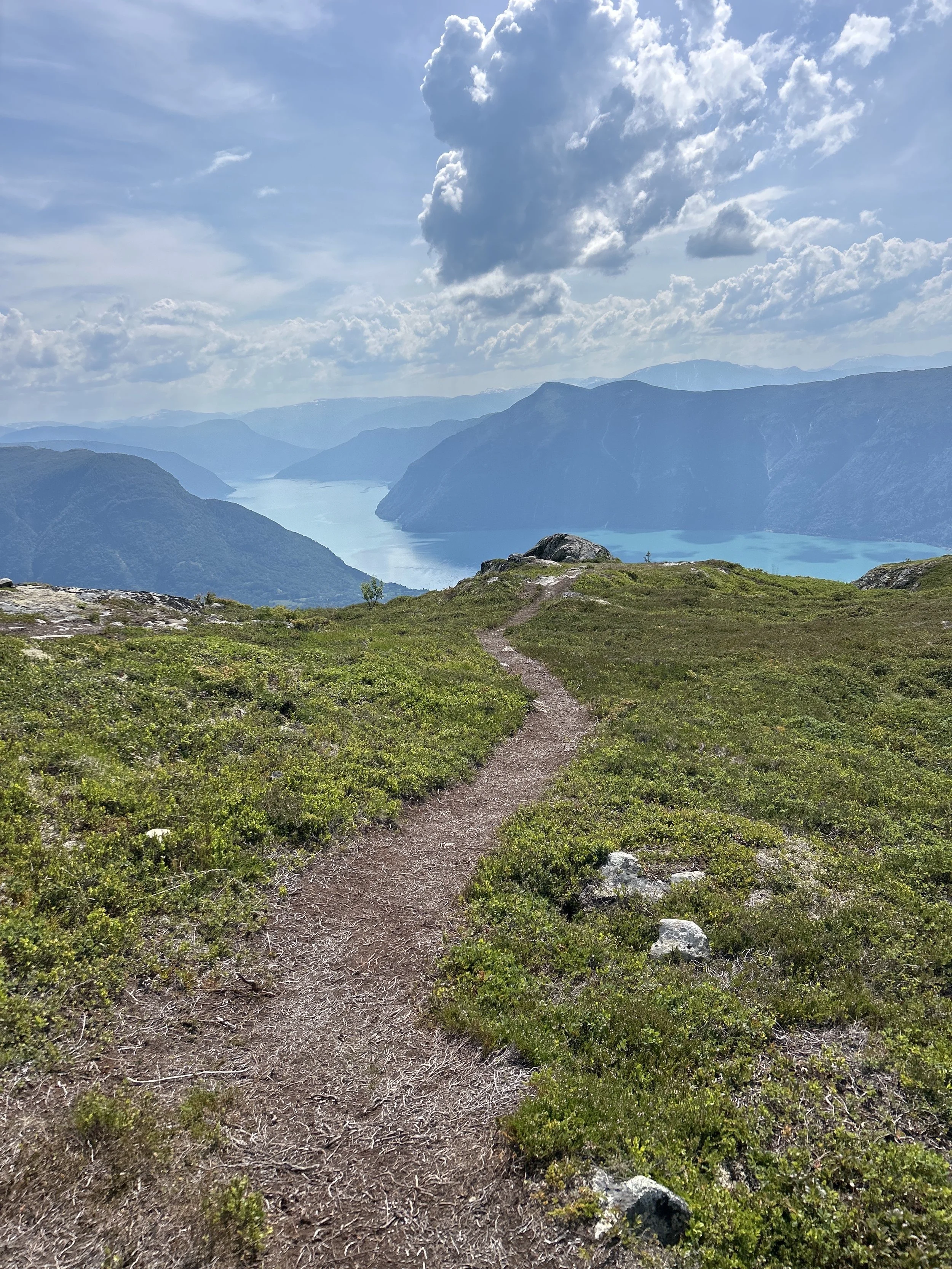 A mountain trail leading to a lake surrounded by mountains under a partly cloudy sky.