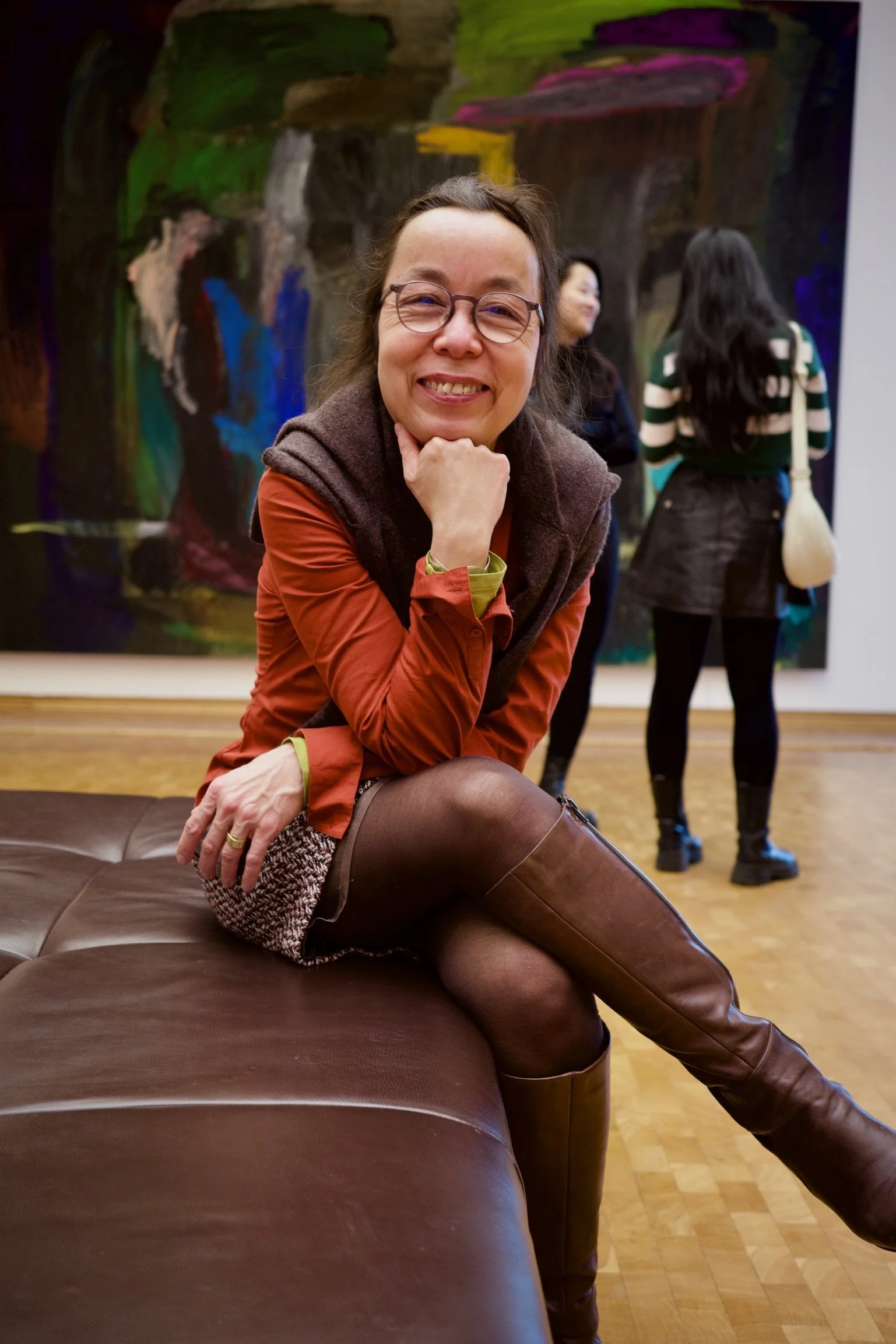 A woman with glasses smiling and sitting on a leather bench, with an abstract painting in the background and several people talking in an art gallery.