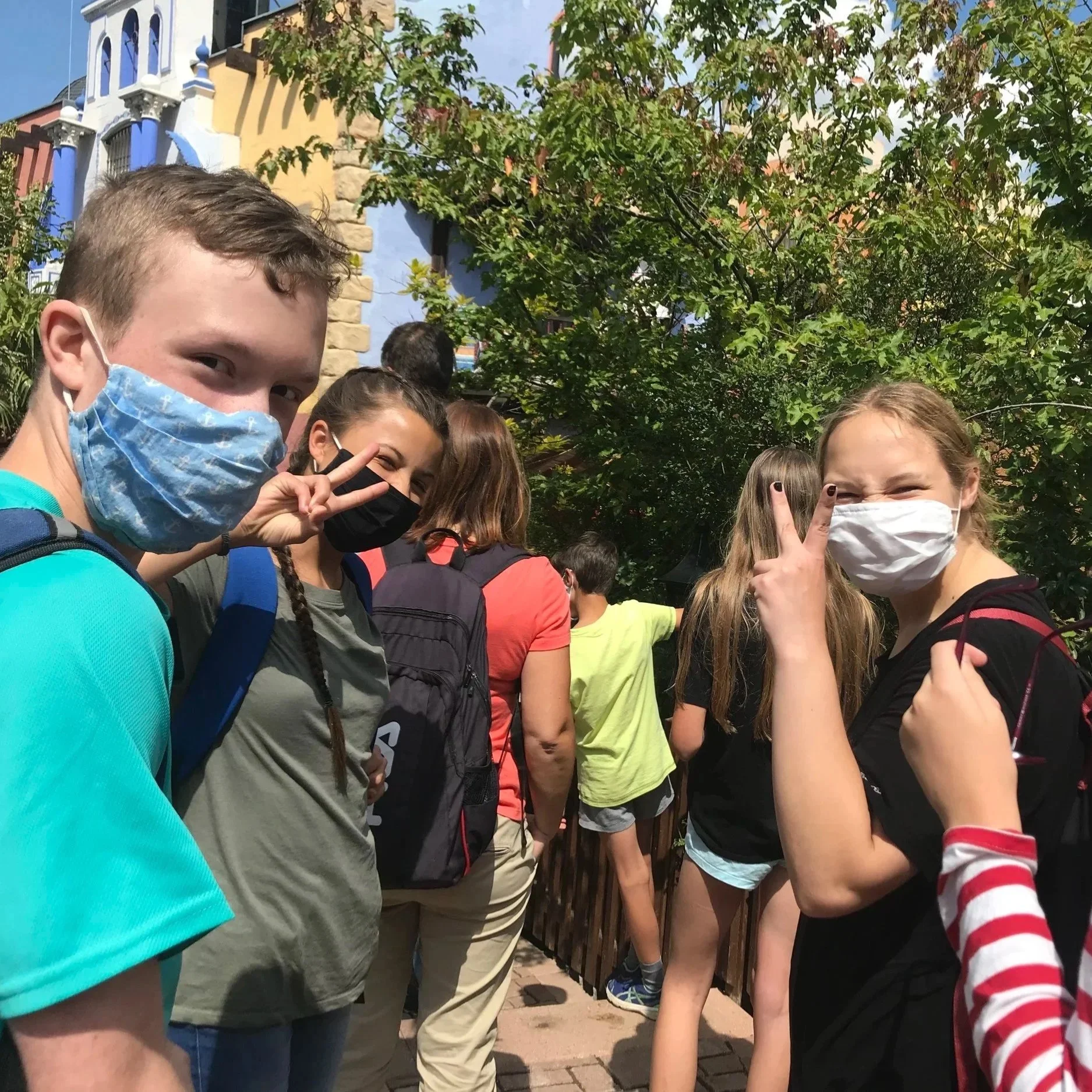 Group of children outdoors wearing face masks, standing in line and making peace signs.