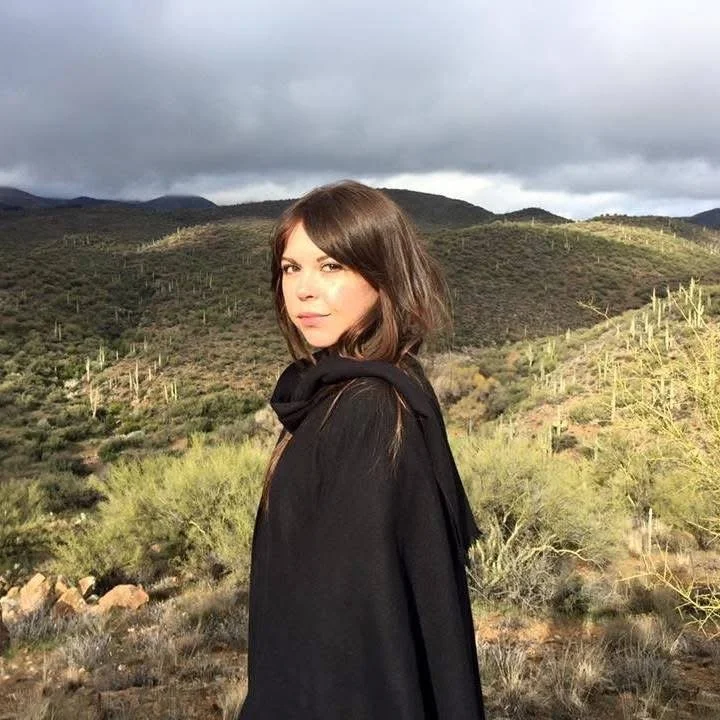 A young woman with shoulder-length brown hair and fair skin standing in a desert landscape with mountains, cacti, and sparse vegetation in the background on a cloudy day.