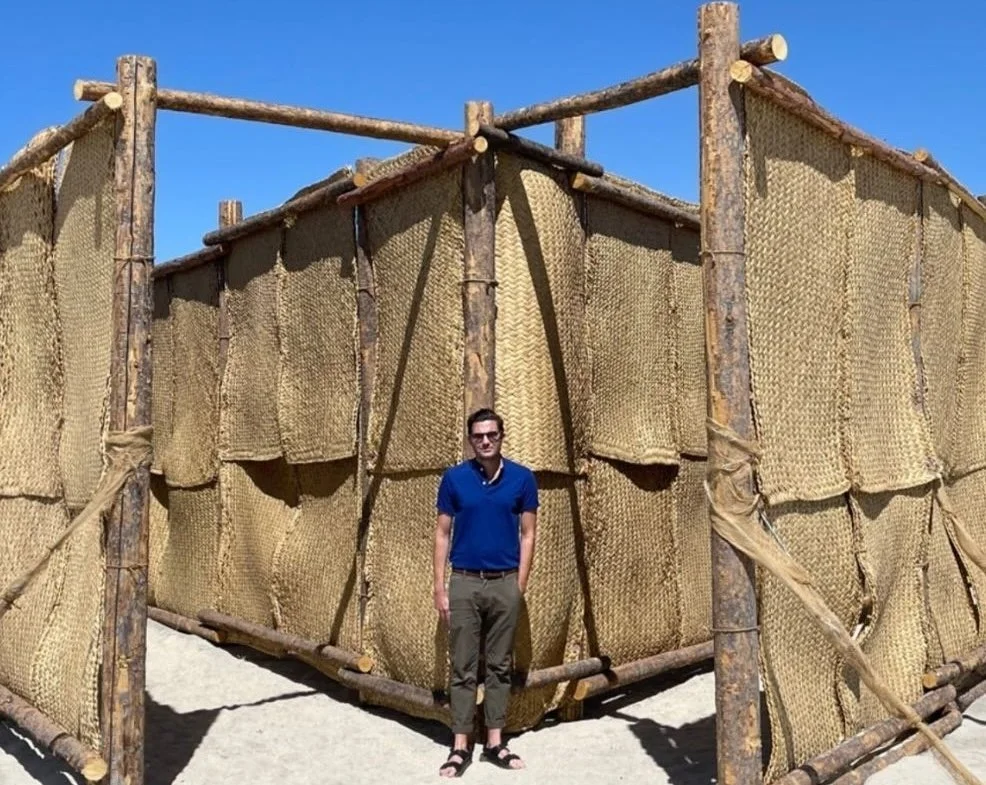A man in sunglasses wearing a blue shirt and khaki pants standing in front of an outdoor structure made of wooden logs and woven material, with a clear blue sky in the background.