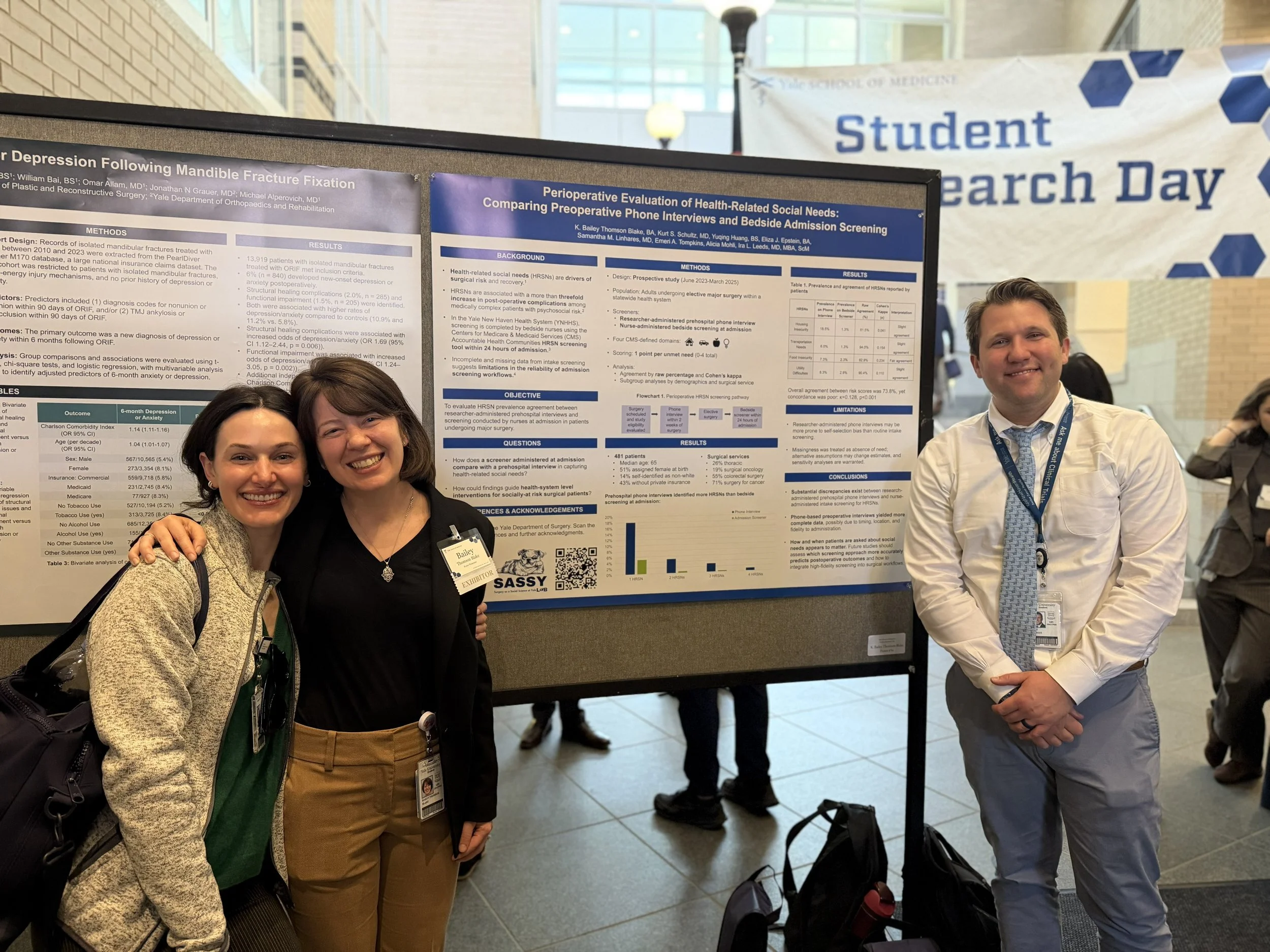 Three people standing in front of a research poster at a conference. The woman on the left has short dark hair and is wearing a beige sweater, a green shirt, and brown pants, and is smiling. The woman in the middle has short dark hair and is wearing a black top and khaki pants, and is smiling. The man on the right has short dark hair and is wearing a white shirt with a blue tie, and is smiling. Behind them, the poster contains scientific data and research information, and there is a background banner that says “Student Research Day.”