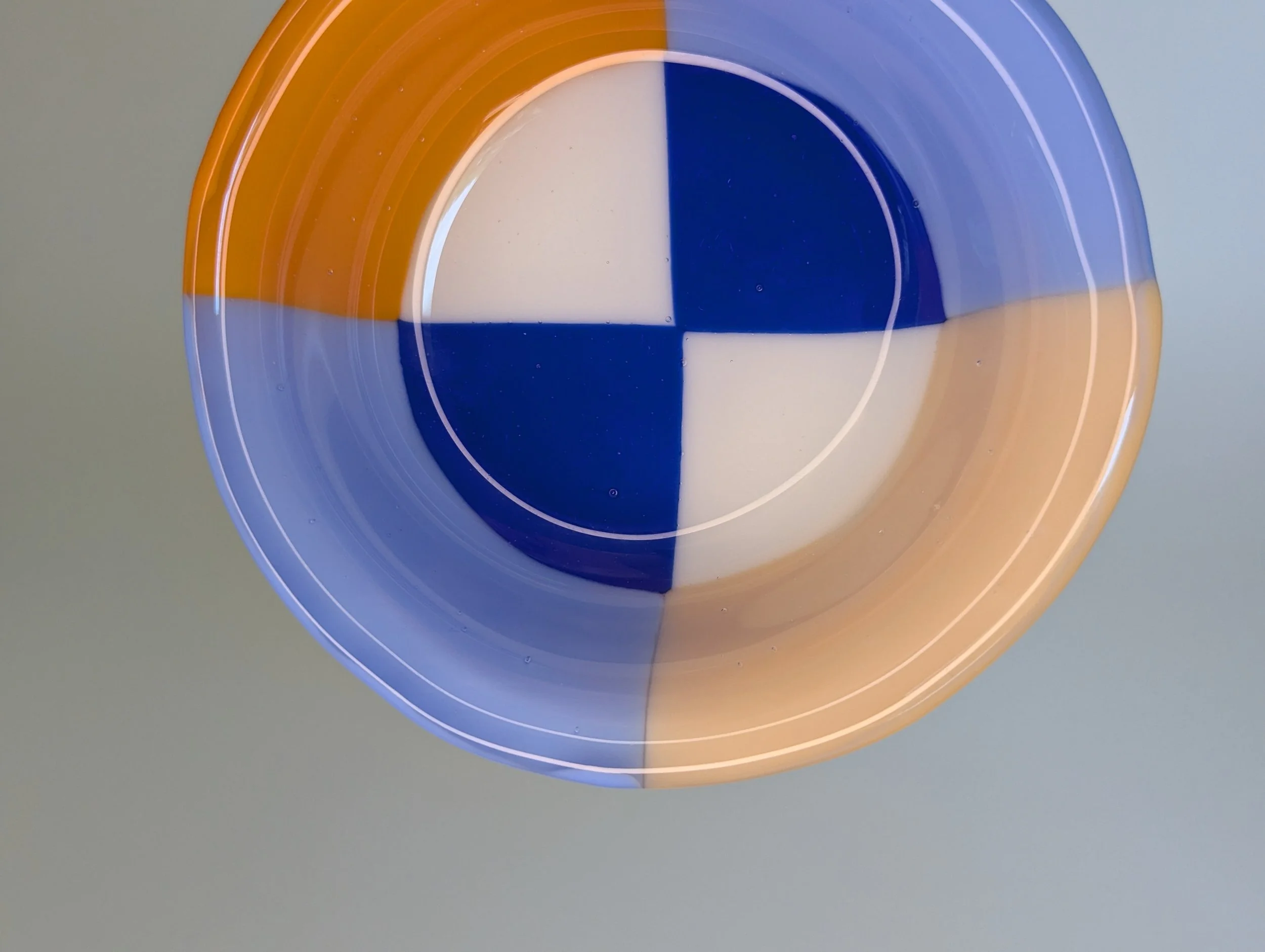 Circular glass bowl with geometric patterns in blue, orange, and white set on a plain light background.