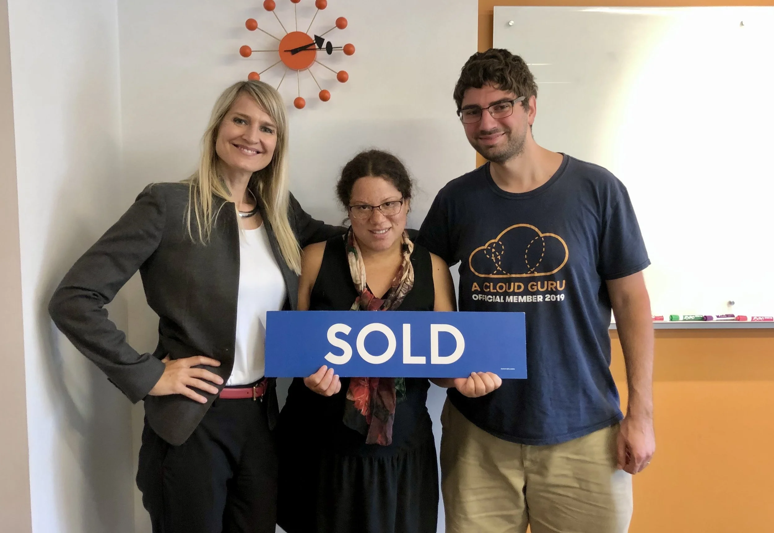 Three people smiling and holding a blue sign that says 'SOLD' in an office. The woman in the middle wears glasses and a scarf. The woman on the left has a black blazer, and the man on the right wears a navy t-shirt with a cloud graphic and the words 
