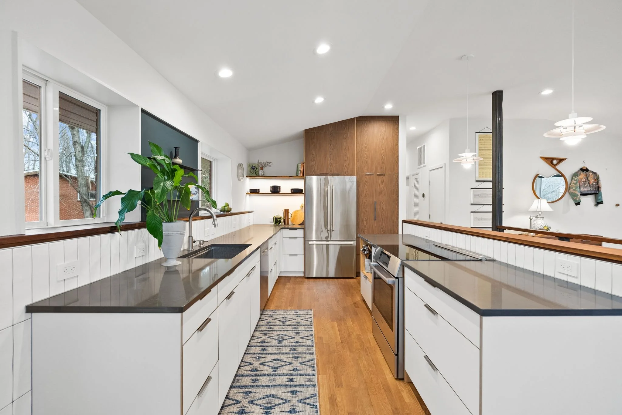 Modern kitchen with white cabinets, wooden accents, stainless steel appliances, black countertops, hardwood floors, and a potted plant on the counter.