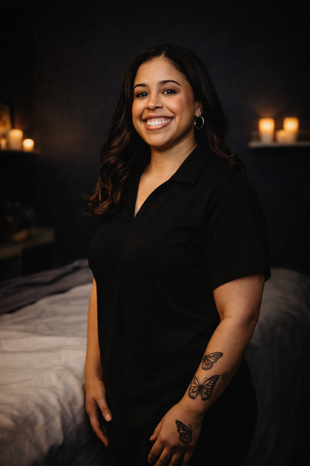A woman with long dark hair and a bright smile, wearing a black shirt, standing in a dimly lit room with candles on a shelf in the background.