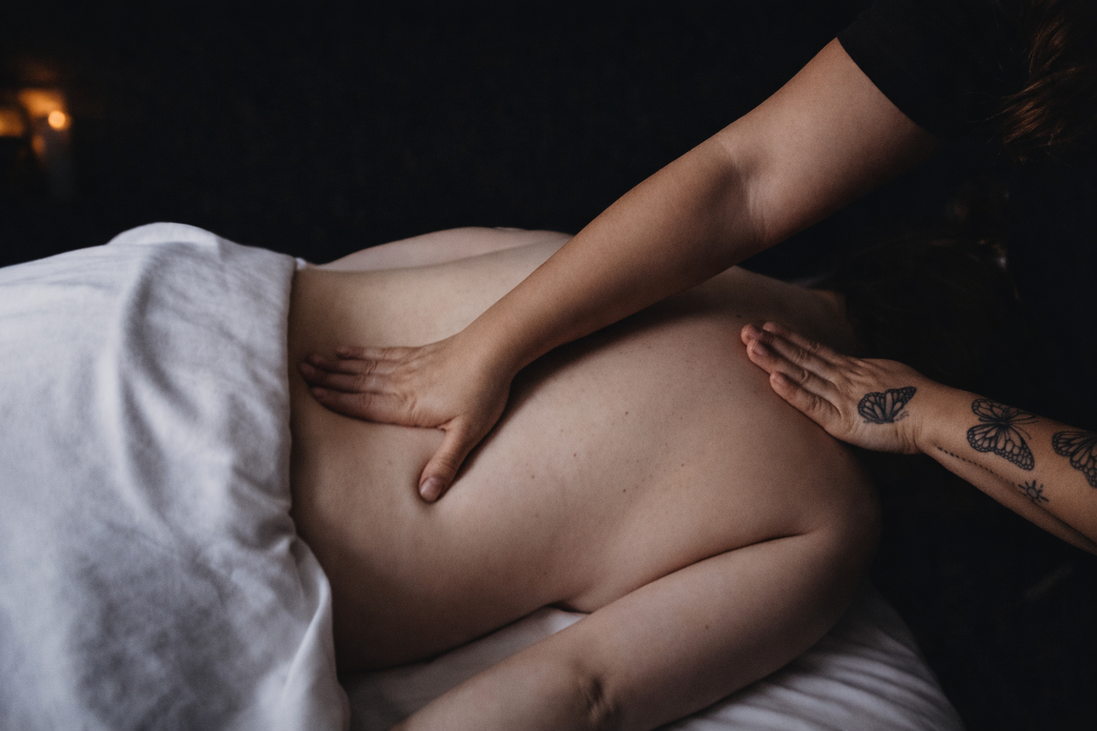 A person receiving a massage on their back from another individual with tattooed arms, with a dark background and soft lighting.