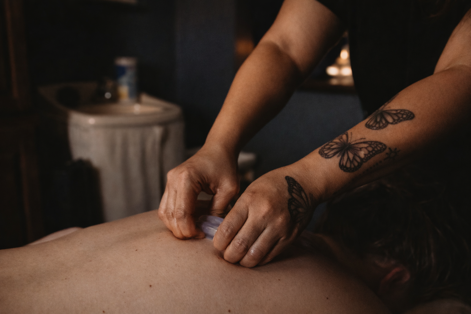 A person with butterfly tattoos on their arm is using a cup or container on a person's back, nearby are dark wooden furniture and a blurred background.
