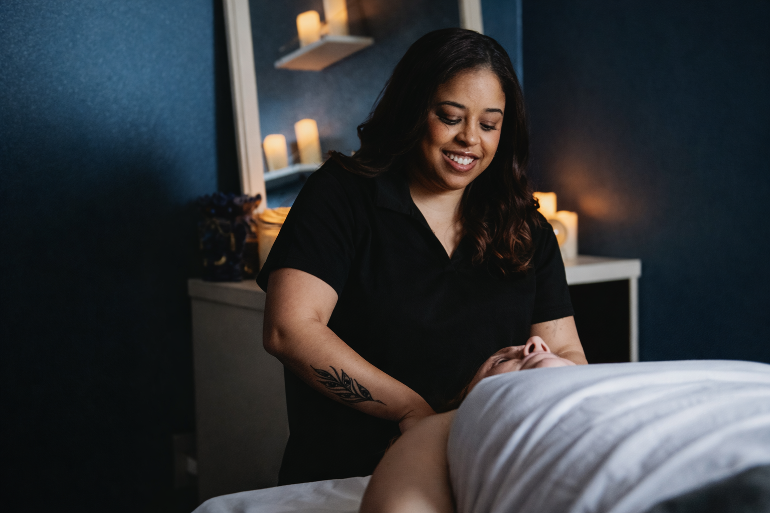 A massage therapist smiling while giving a massage to a client lying face up in a dimly lit room with candles in the background.