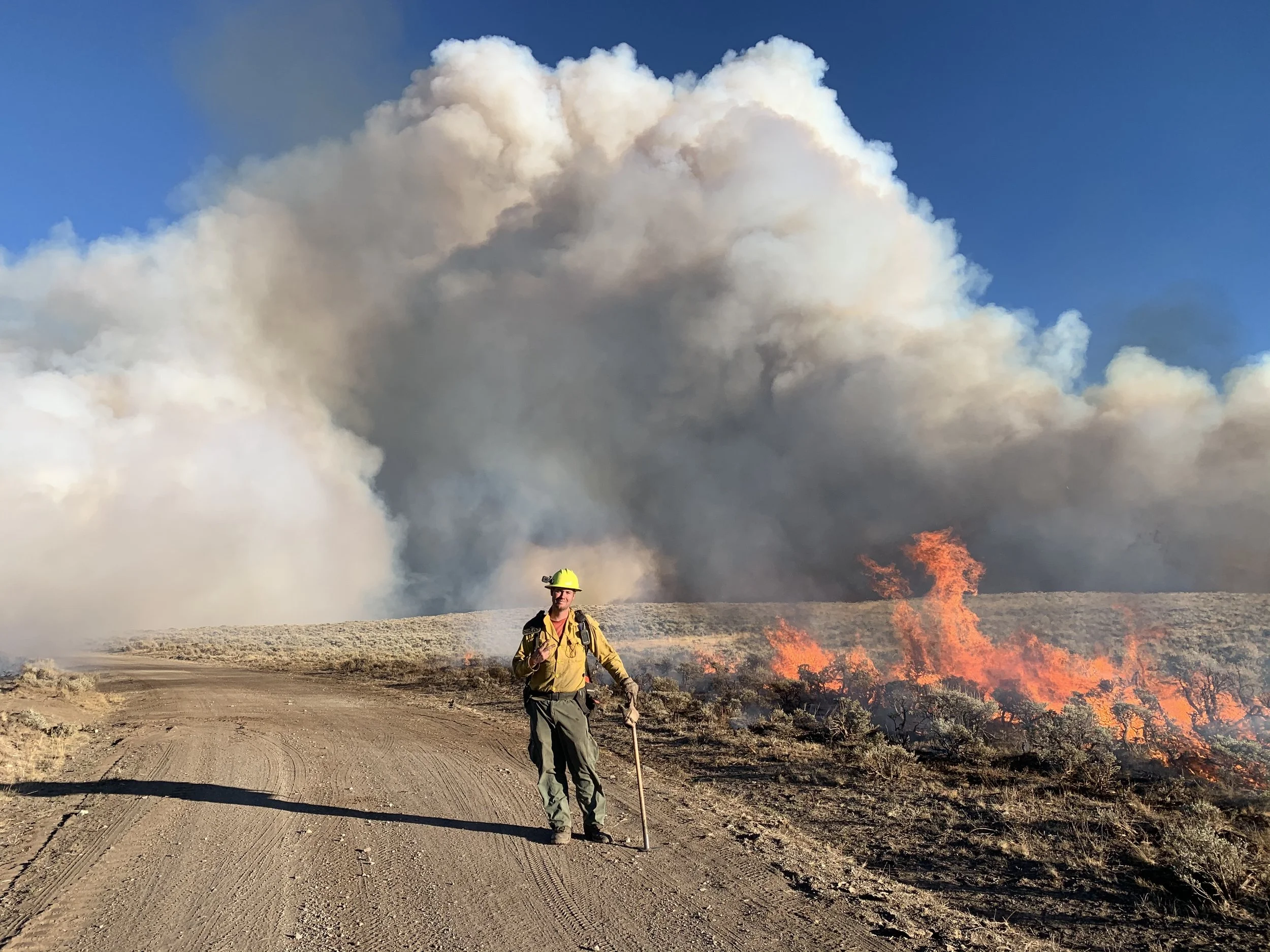 A firefighter in yellow gear walking on a dirt road, with a wildfire burning along the side and large smoke clouds in the background.