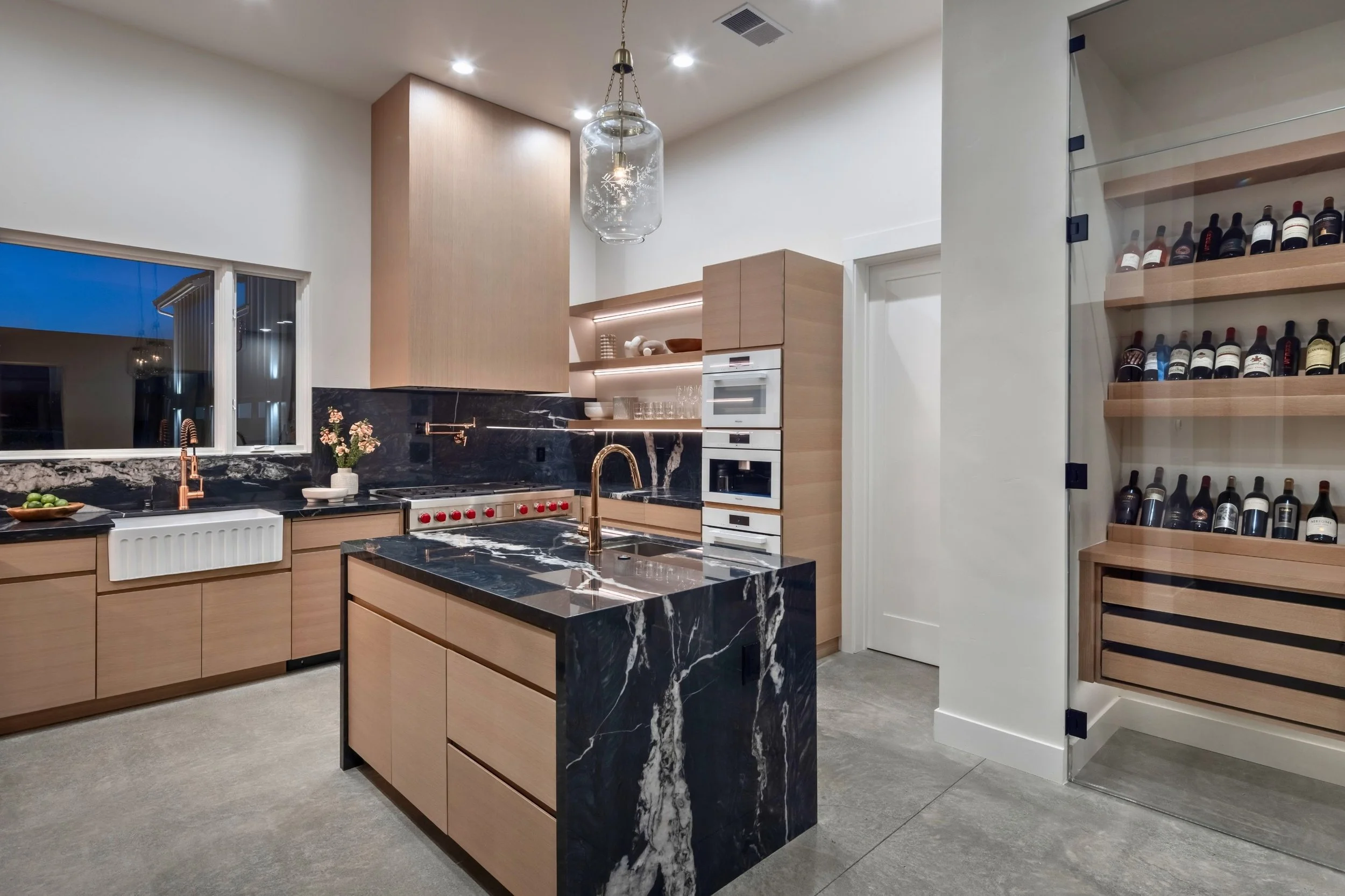 Modern kitchen with black marble island, light wood cabinetry, and open shelving. Features a stainless steel stove, gold faucet, and a wine display on the right. Bright lighting and a window show evening outside.