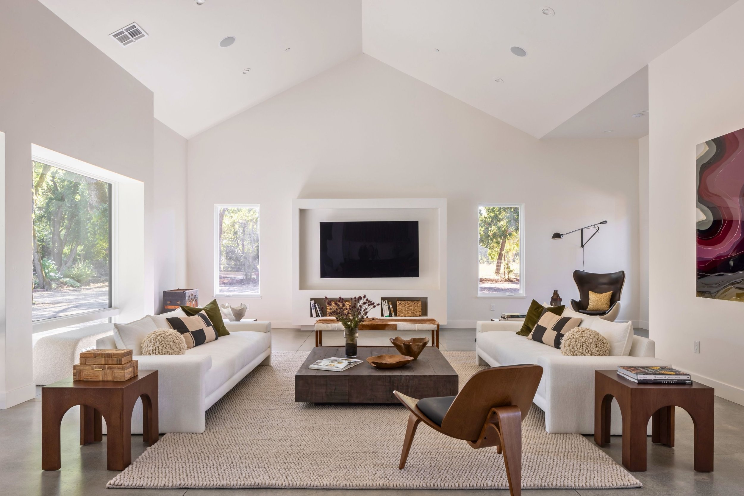 Bright living room with white walls, vaulted ceiling, large windows, a flat-screen TV, two white sofas with pillows, a wooden coffee table, a decorative bench, a leather chair, and contemporary artwork.