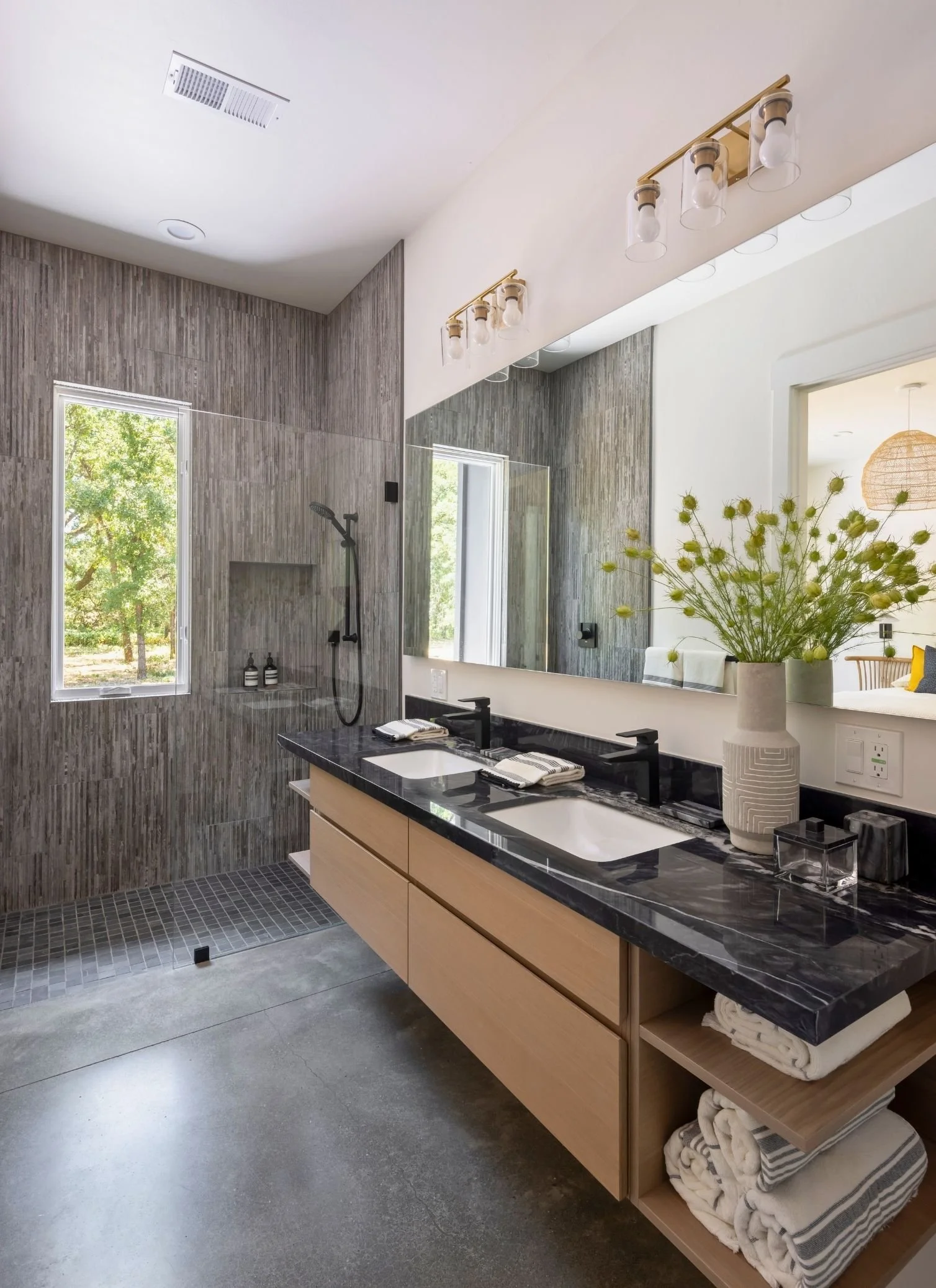 Modern bathroom with dual sinks, black marble countertop, wooden cabinetry, large mirror, and a shower area with a window and gray tiles