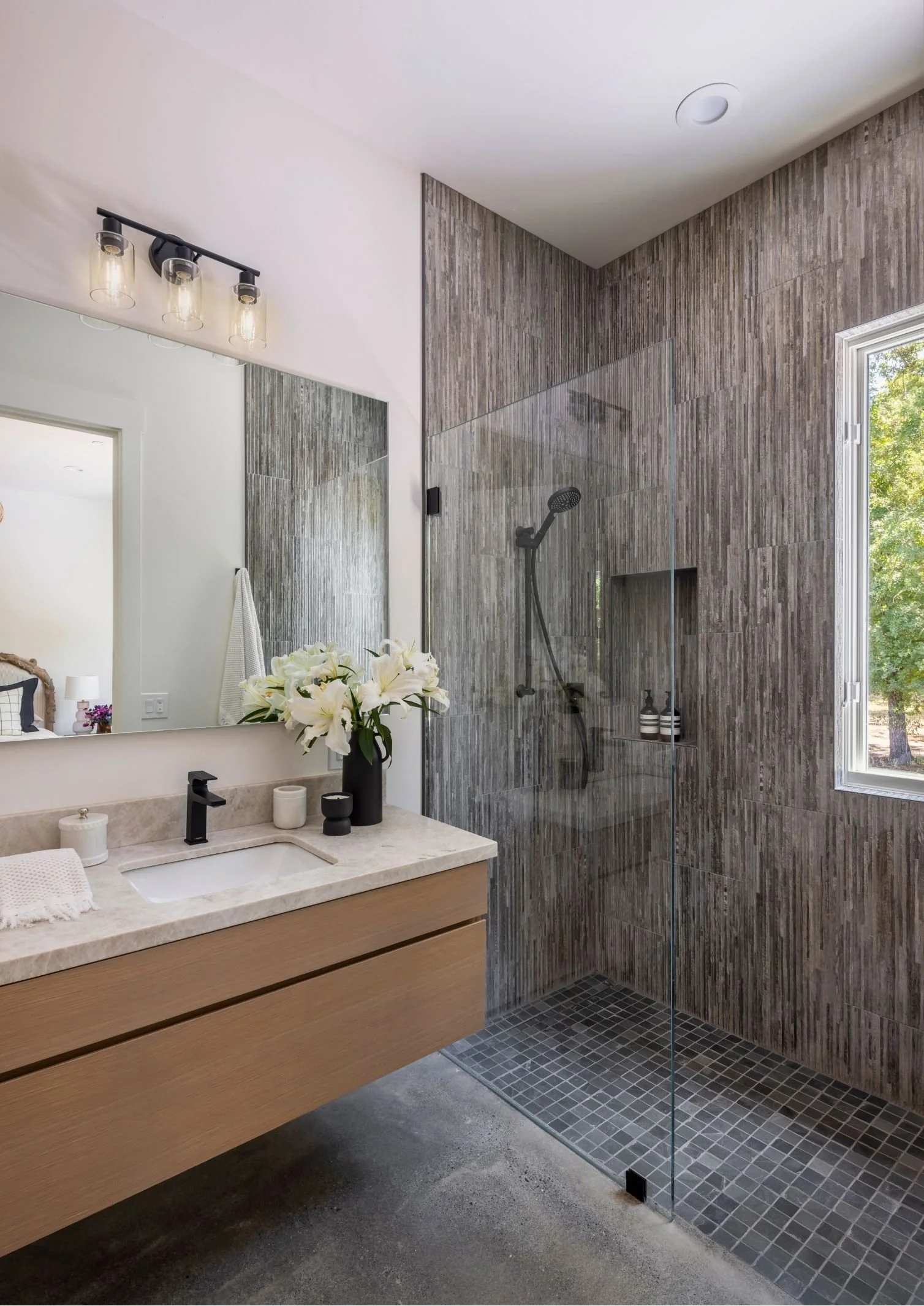 Modern bathroom with a wooden vanity, white sink, black faucet, mirror, flower vase, and toiletries; corner shower with glass partition, black showerhead, niche with bottles, gray tile walls, and window showing green trees outside.