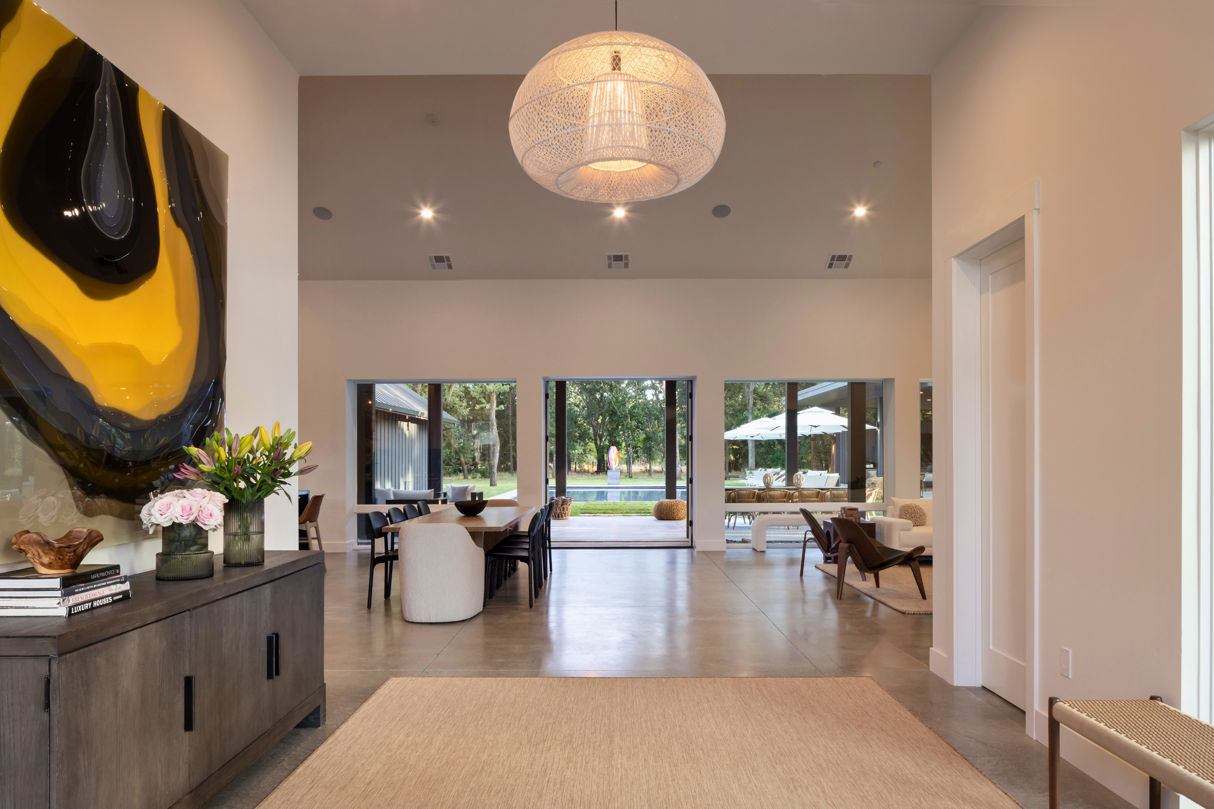 Open-concept living and dining area with large windows overlooking a backyard pool, modern furniture, a statement artwork on the wall, and a woven pendant light hanging from the ceiling.