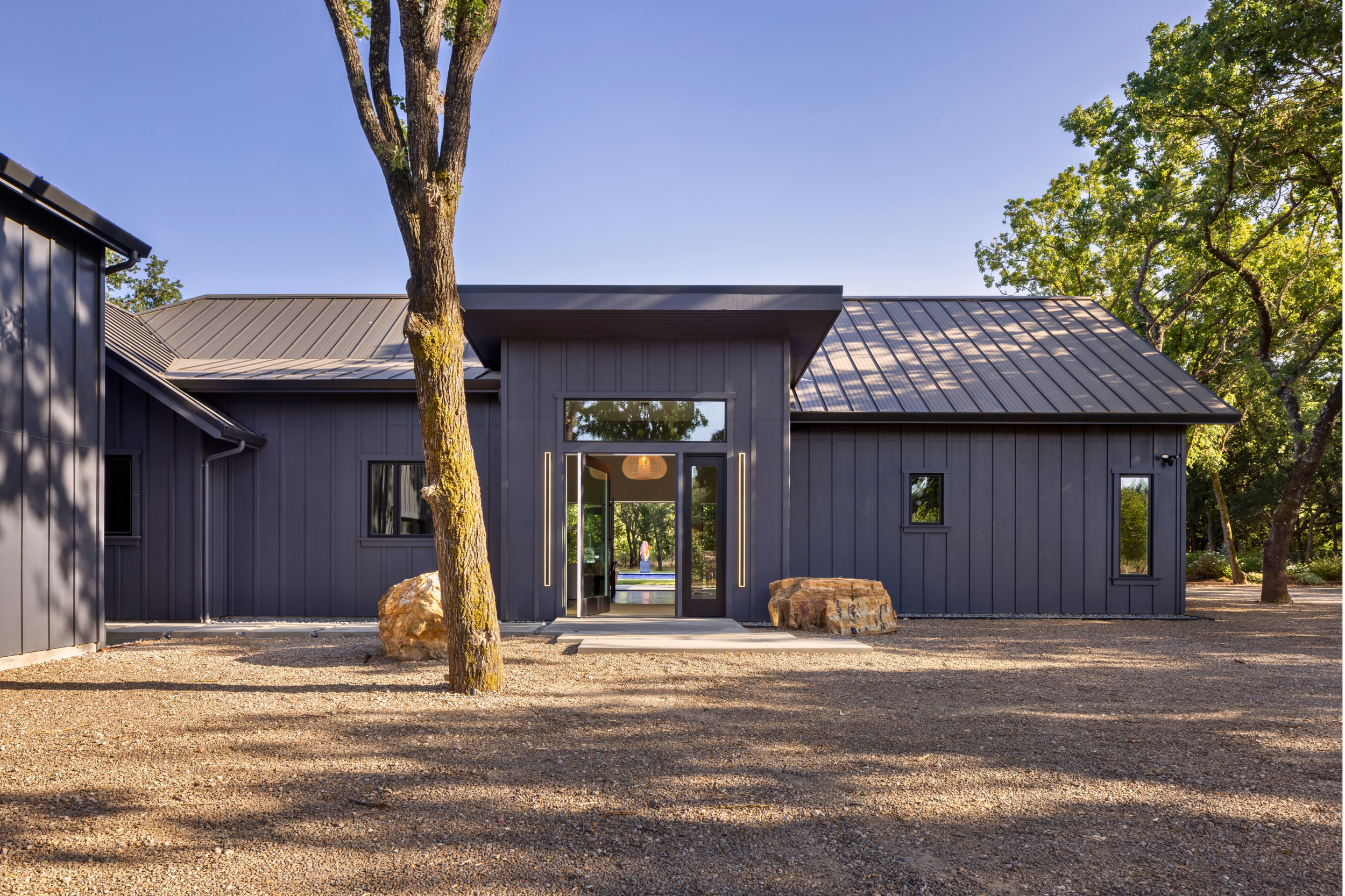 Modern dark gray building with metal roof, surrounded by trees, with large rocks and a gravel ground in front.