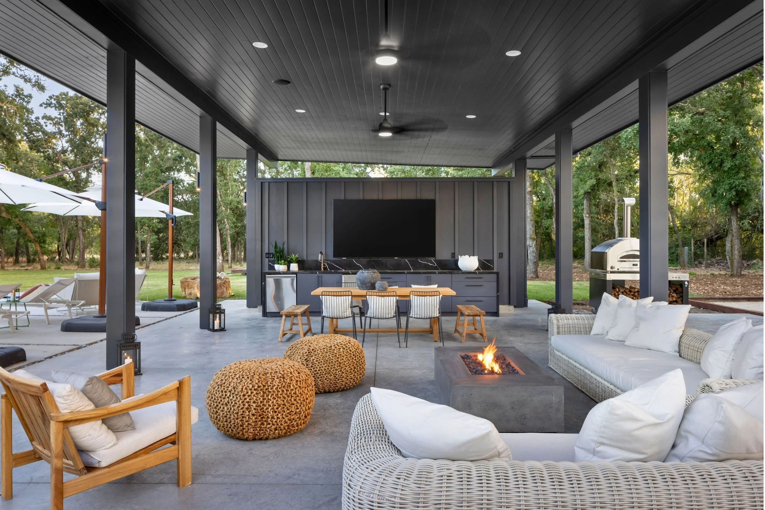 Covered outdoor patio area with a modern seating arrangement, a firepit, a TV, and an outdoor kitchen space surrounded by trees.
