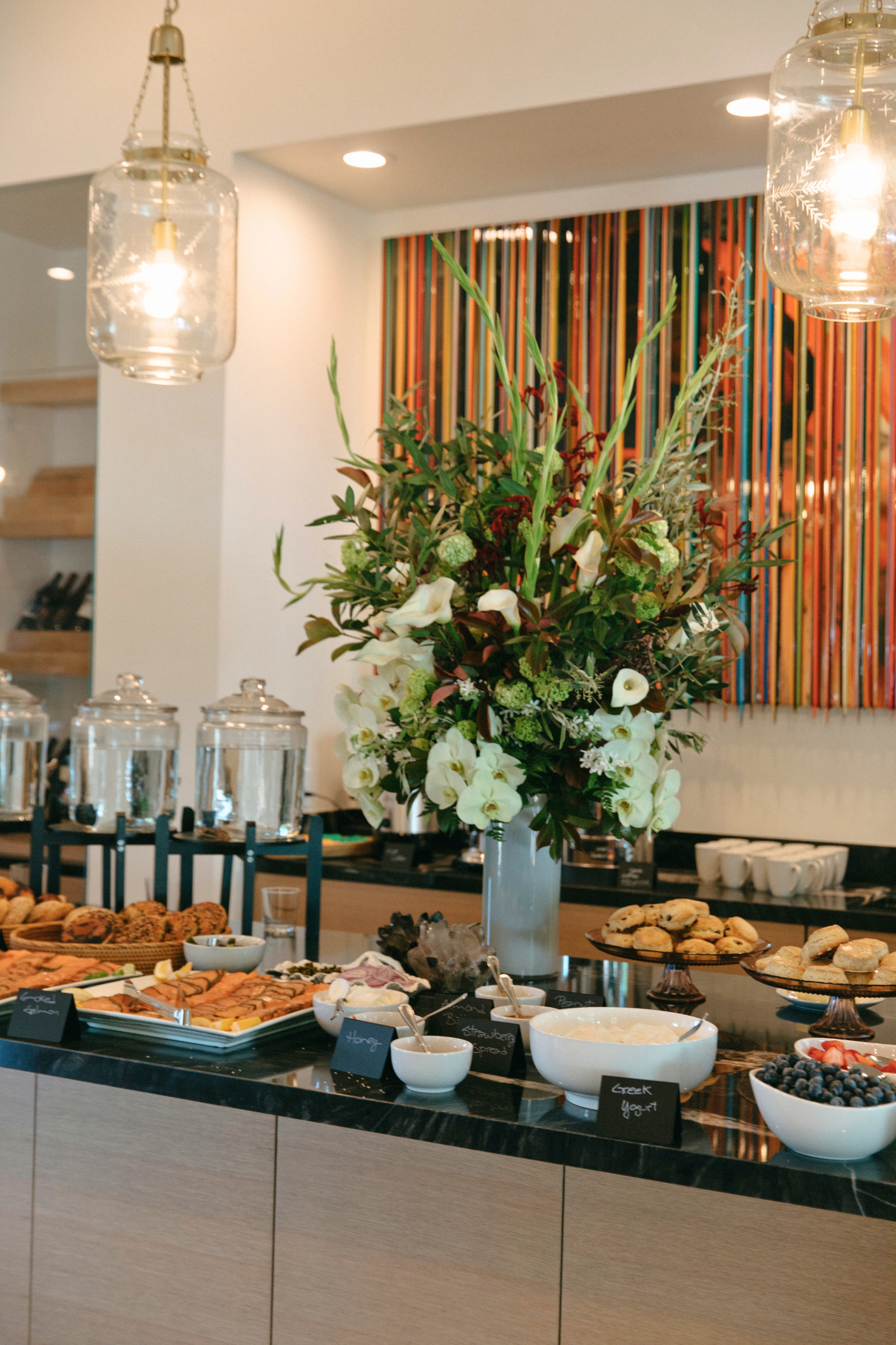 A buffet table with a large flower arrangement, bowls of blueberries and Greek yogurt, cookies, and small bowls of honey, strawberry preserve, and other toppings.