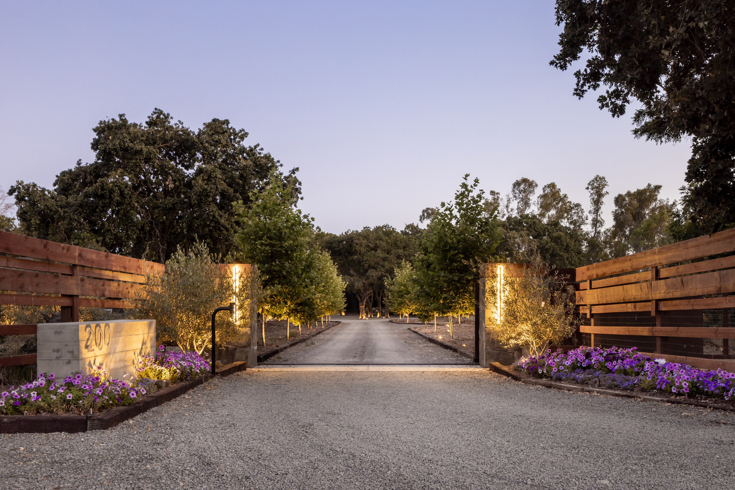 A gated entrance to a driveway with landscaped flower beds on either side, illuminated by outdoor lighting, leading into a wooded area at dusk.