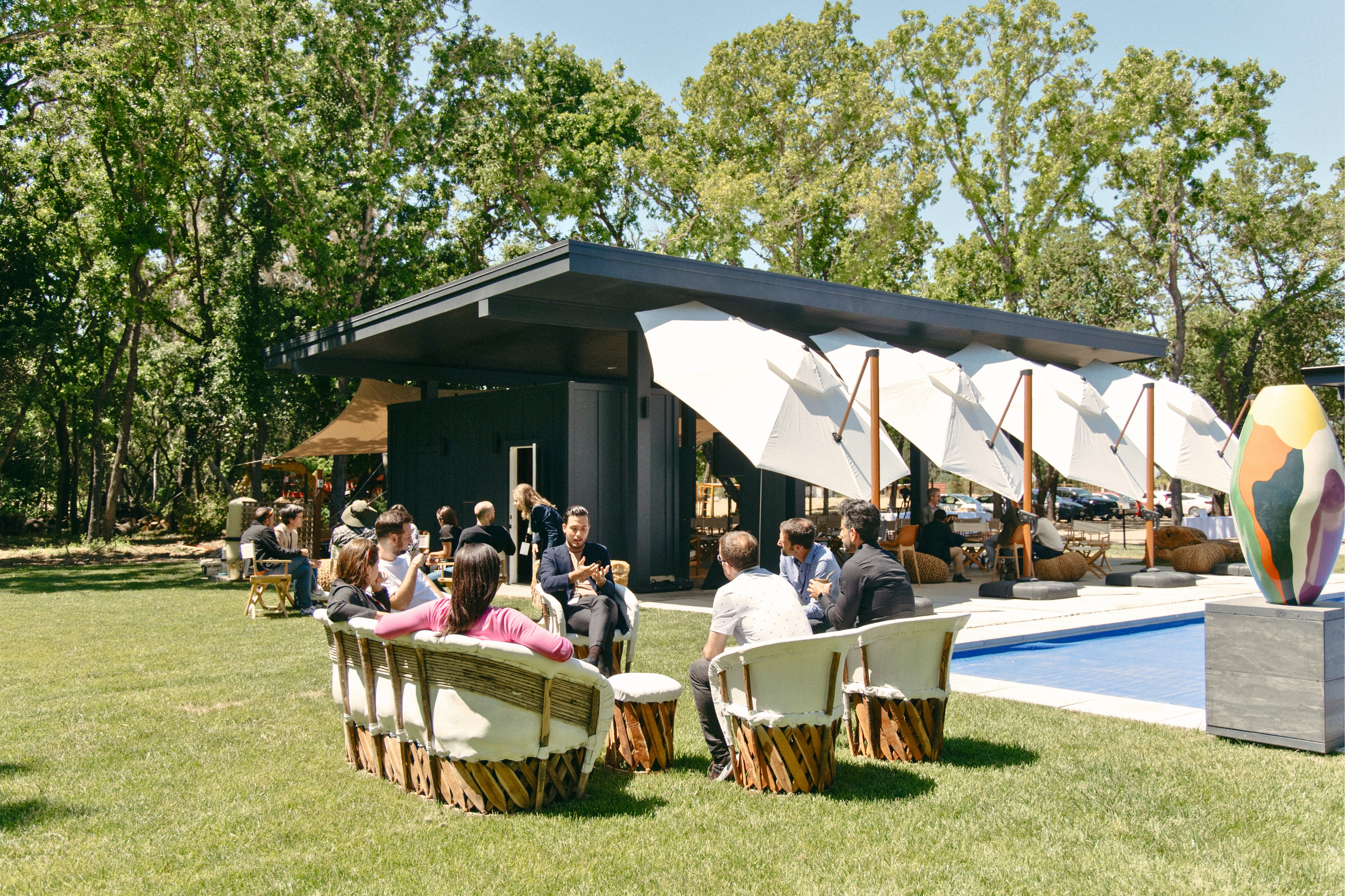 People seated outdoors near a modern dark-colored building with large white umbrellas, a swimming pool, and decorative plants, surrounded by trees on a sunny day.
