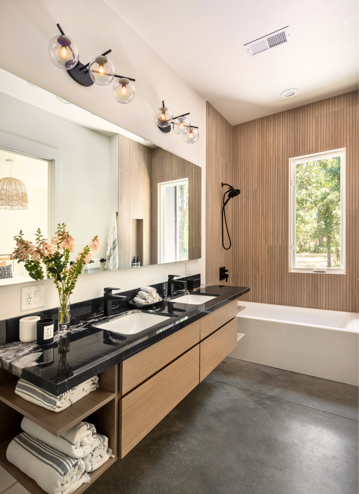 Modern bathroom with double sink vanity, black marble countertop, potted flowers, wooden cabinetry, large mirror, black fixtures, wooden accent wall, bathtub by a window, wood paneling, and a black showerhead.