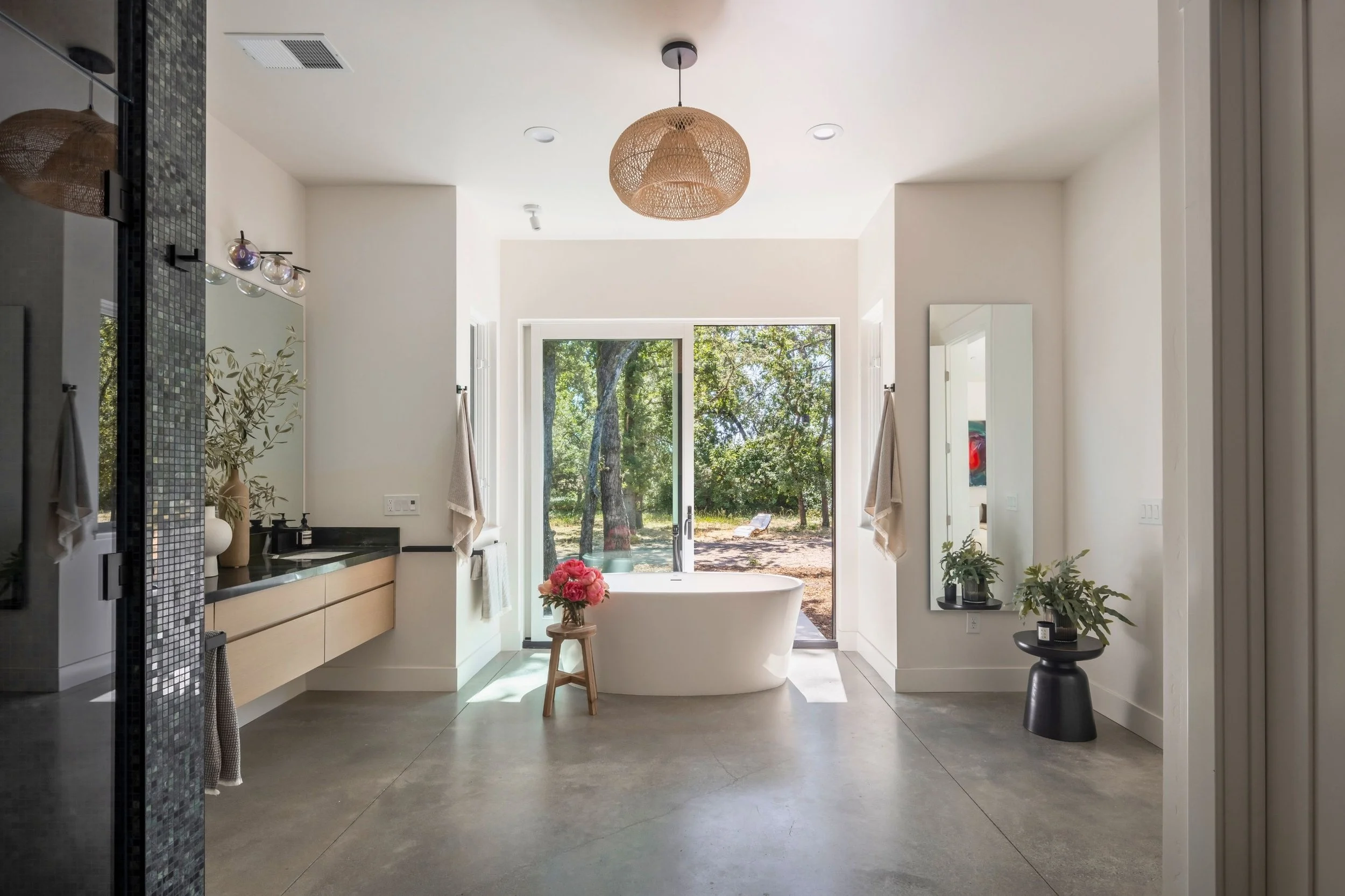 Modern bathroom with a freestanding bathtub near large glass sliding doors to outdoor greenery, a vanity with a mirror and soap dispenser, potted plants, and a woven ceiling light.
