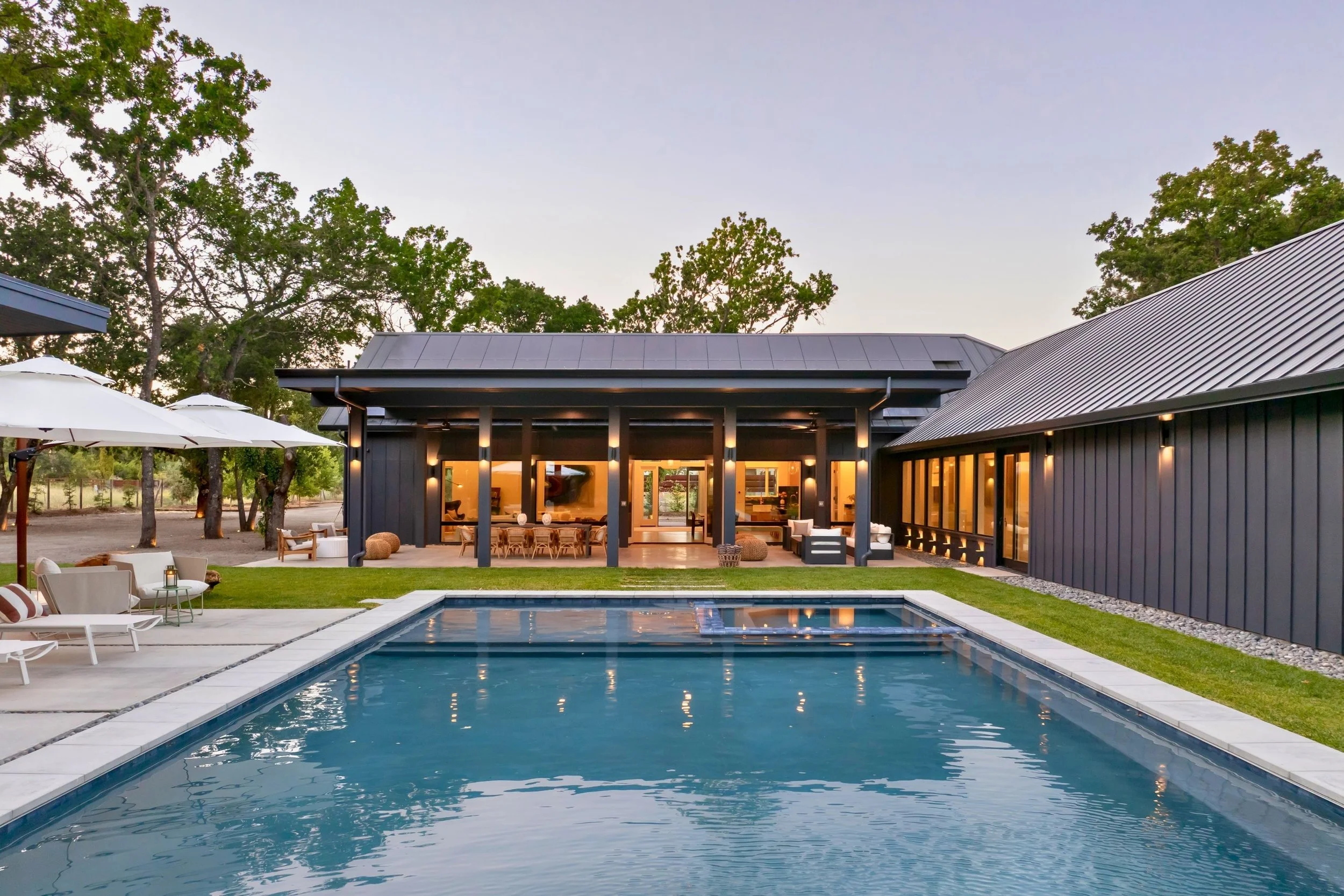 Modern backyard with a rectangular swimming pool, outdoor lounge chairs, patio umbrellas, and a black house with large glass doors and windows, surrounded by trees.