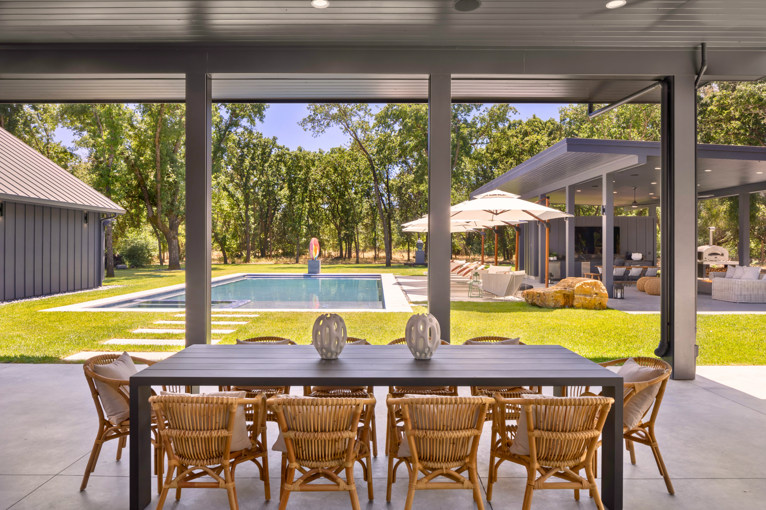 View through a covered patio area to a backyard with a pool, outdoor seating, umbrellas, and trees.
