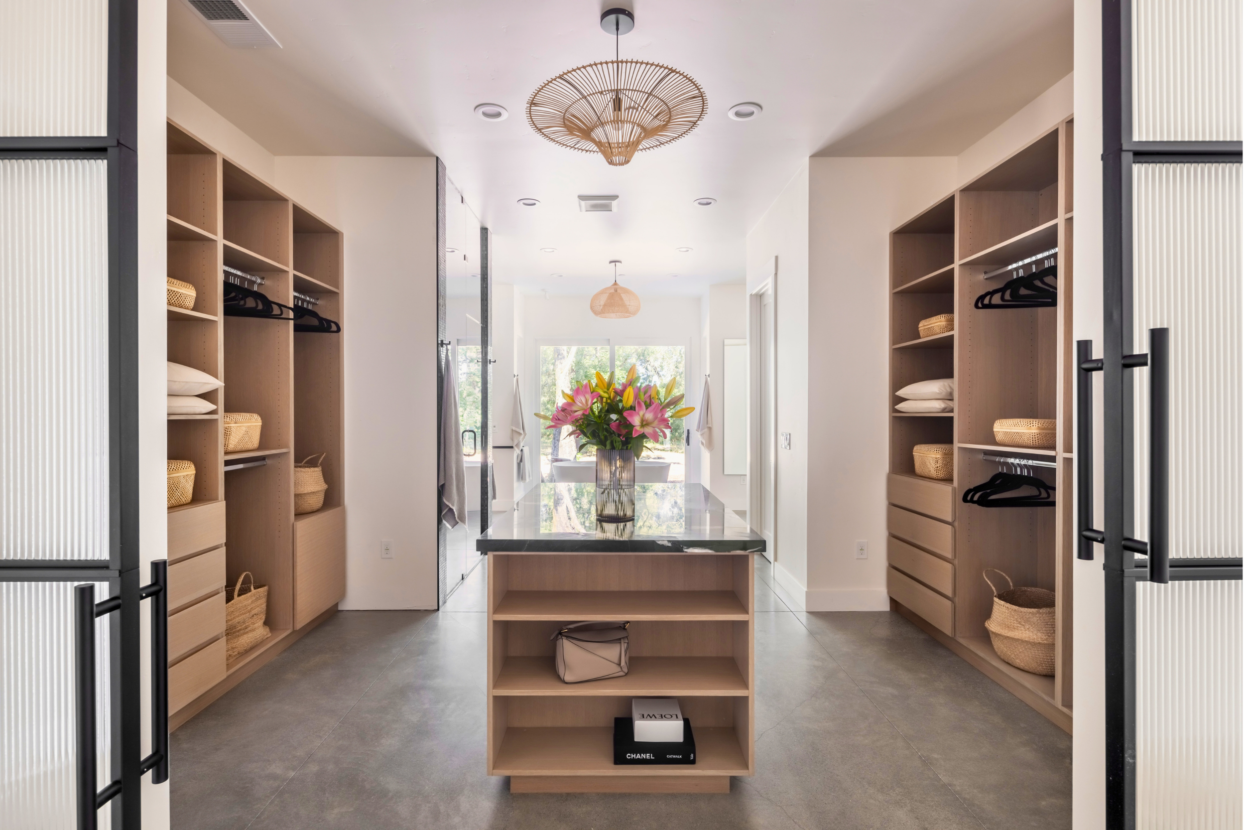Modern walk-in closet with built-in wooden shelves, a black marble-topped island with a pink lily bouquet, and a view of a bright outdoor area through large windows.