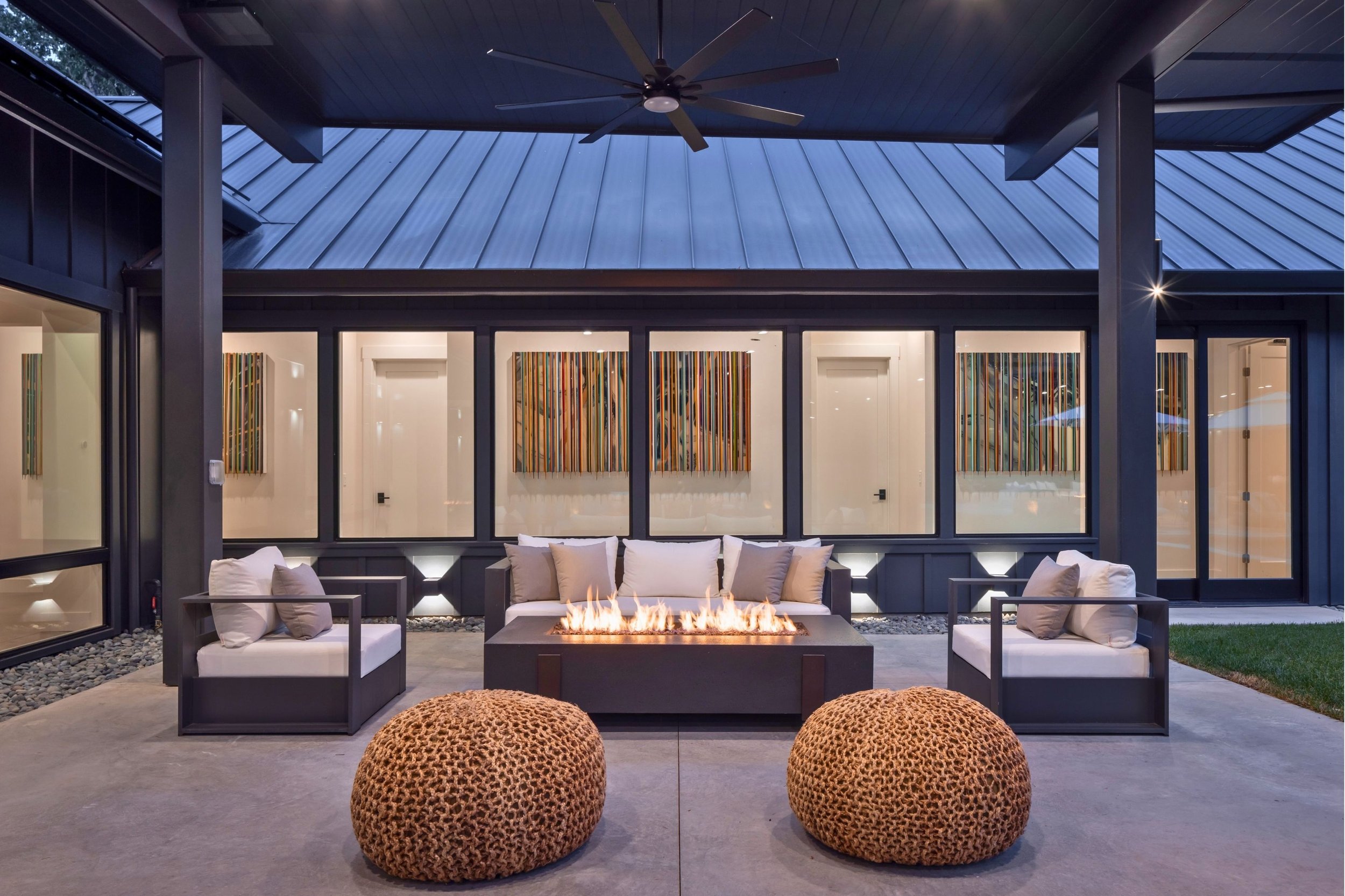 Modern outdoor patio with firepit, cushioned seating, and abstract art on windows, under a dark metal roof