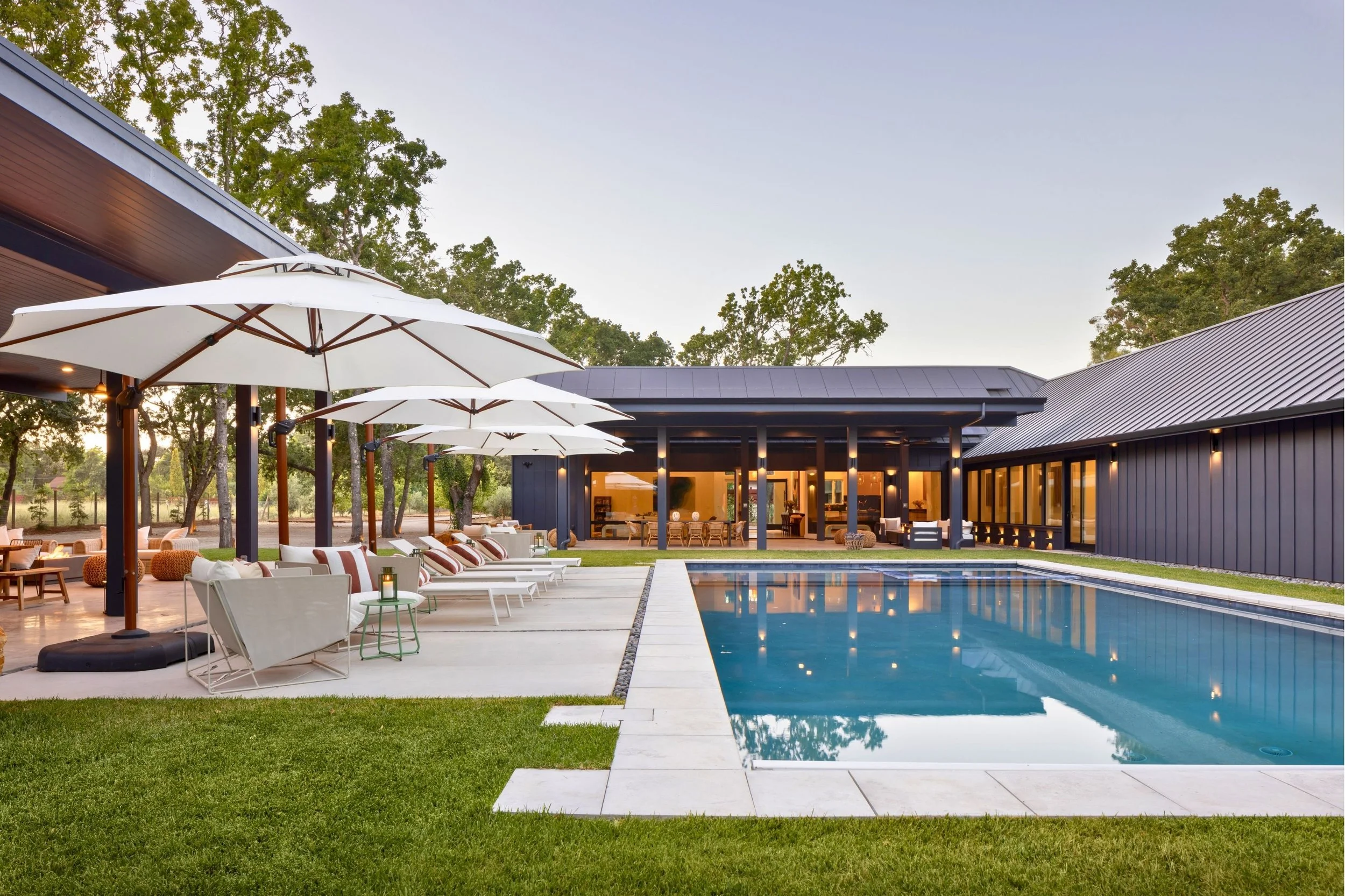 Modern backyard with outdoor pool, lounge chairs under large white umbrellas, and a contemporary house with large windows at dusk.