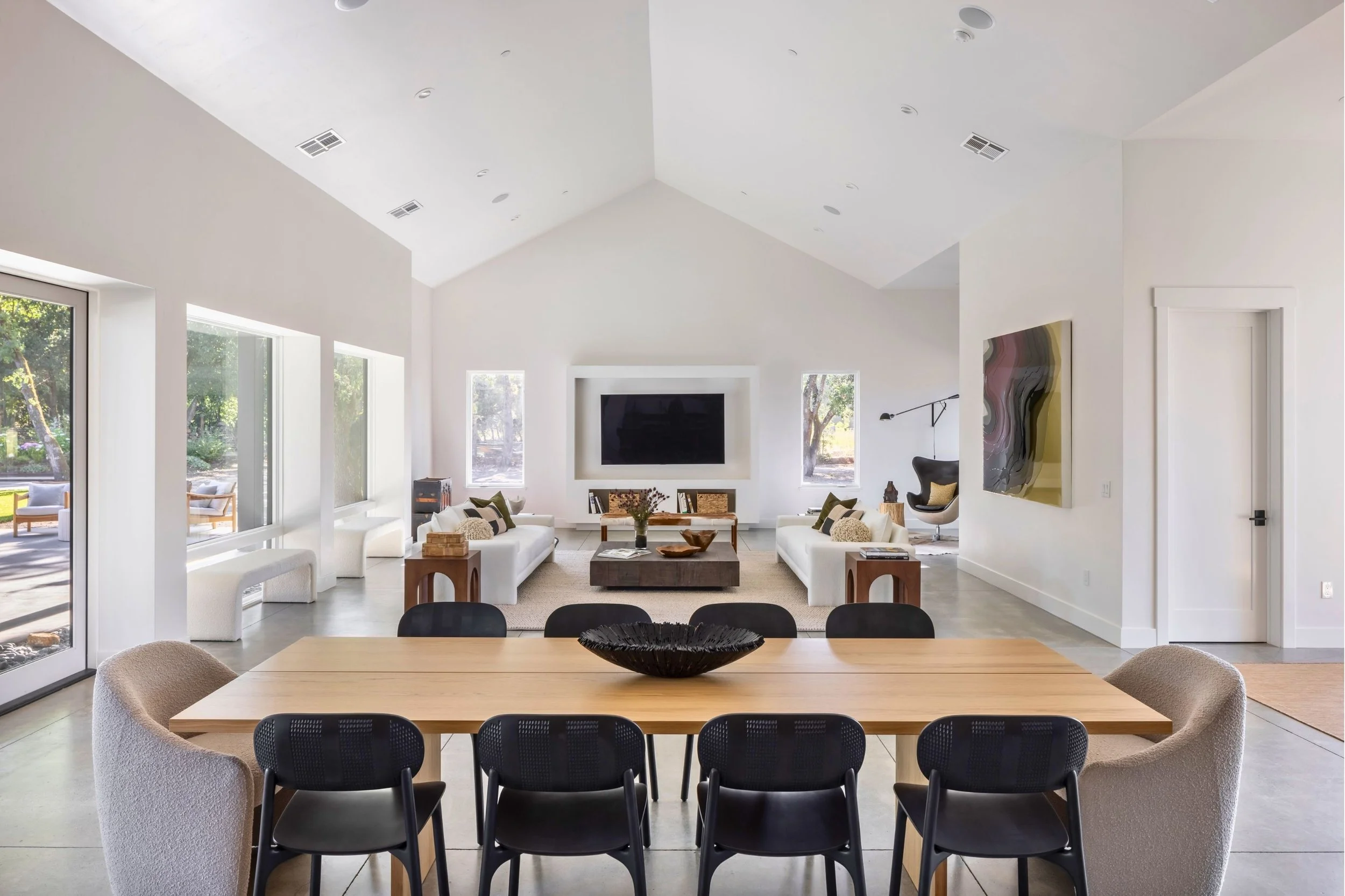 Open-plan living and dining room with large white sofas, a wooden coffee table, and a flat-screen TV on the wall, with a dining table and chairs in the foreground and large windows providing natural light.