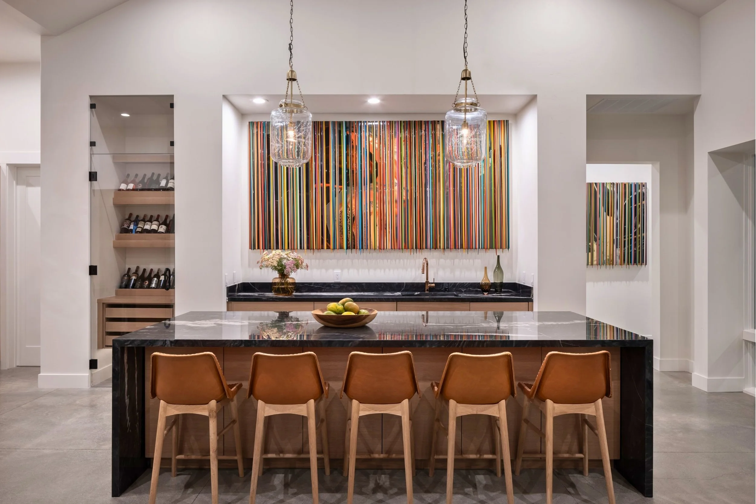 Modern kitchen with a marble island, four chairs, artwork, and wine bottles on shelves.