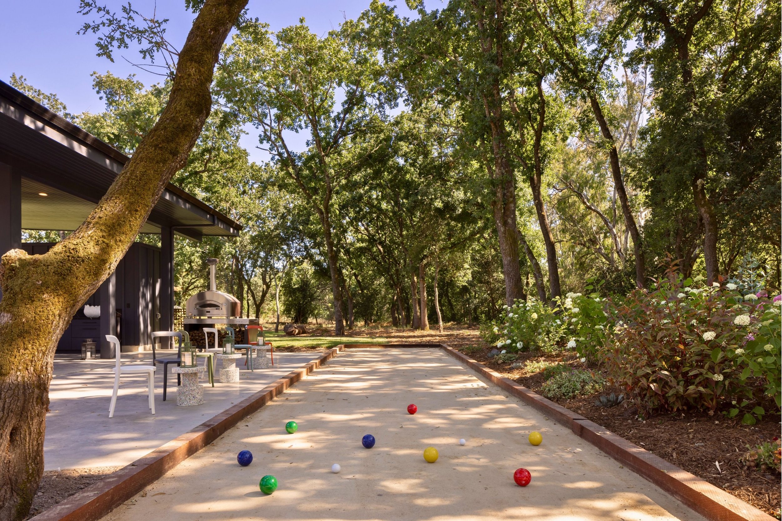 A backyard outdoor bocce court with colorful bocce balls and a shaded patio area with chairs and a pizza oven, surrounded by trees and gardens.