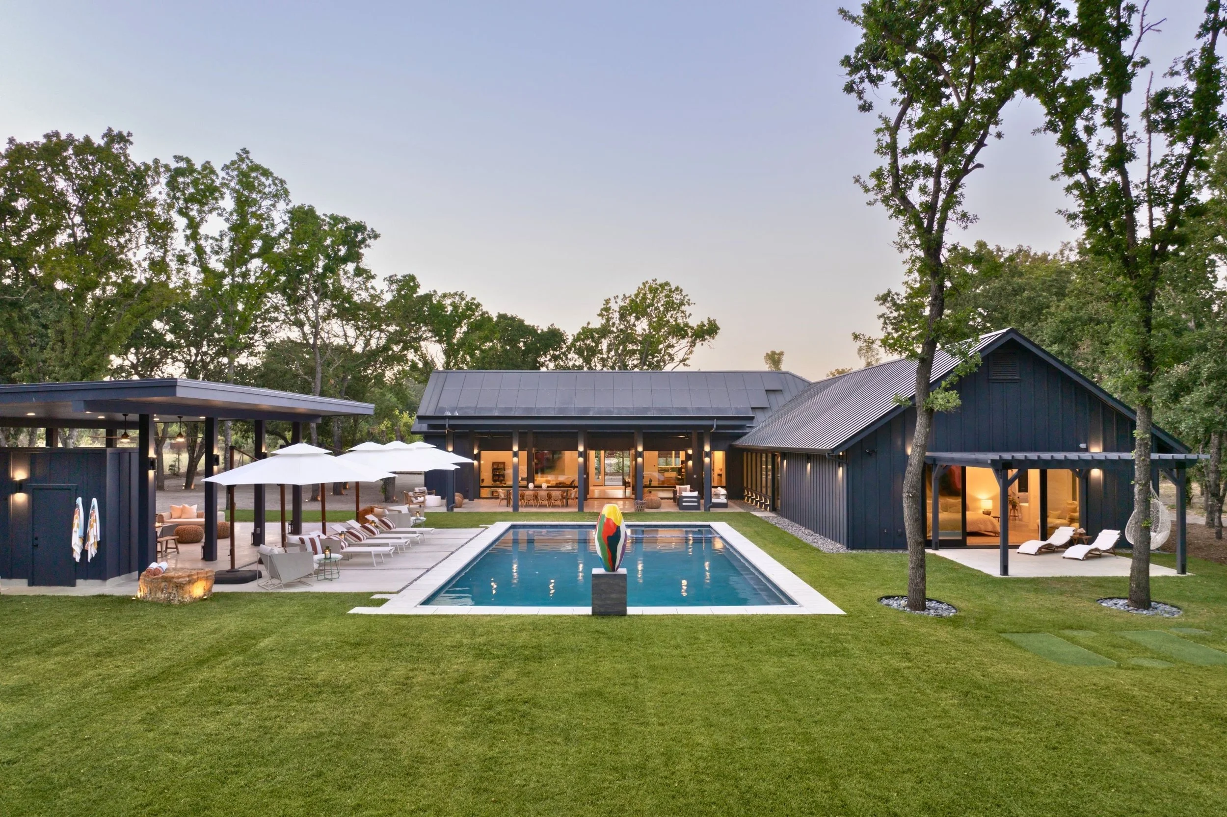 Modern backyard with swimming pool, lounge chairs, umbrellas, and black buildings surrounded by green trees and grass at sunset.