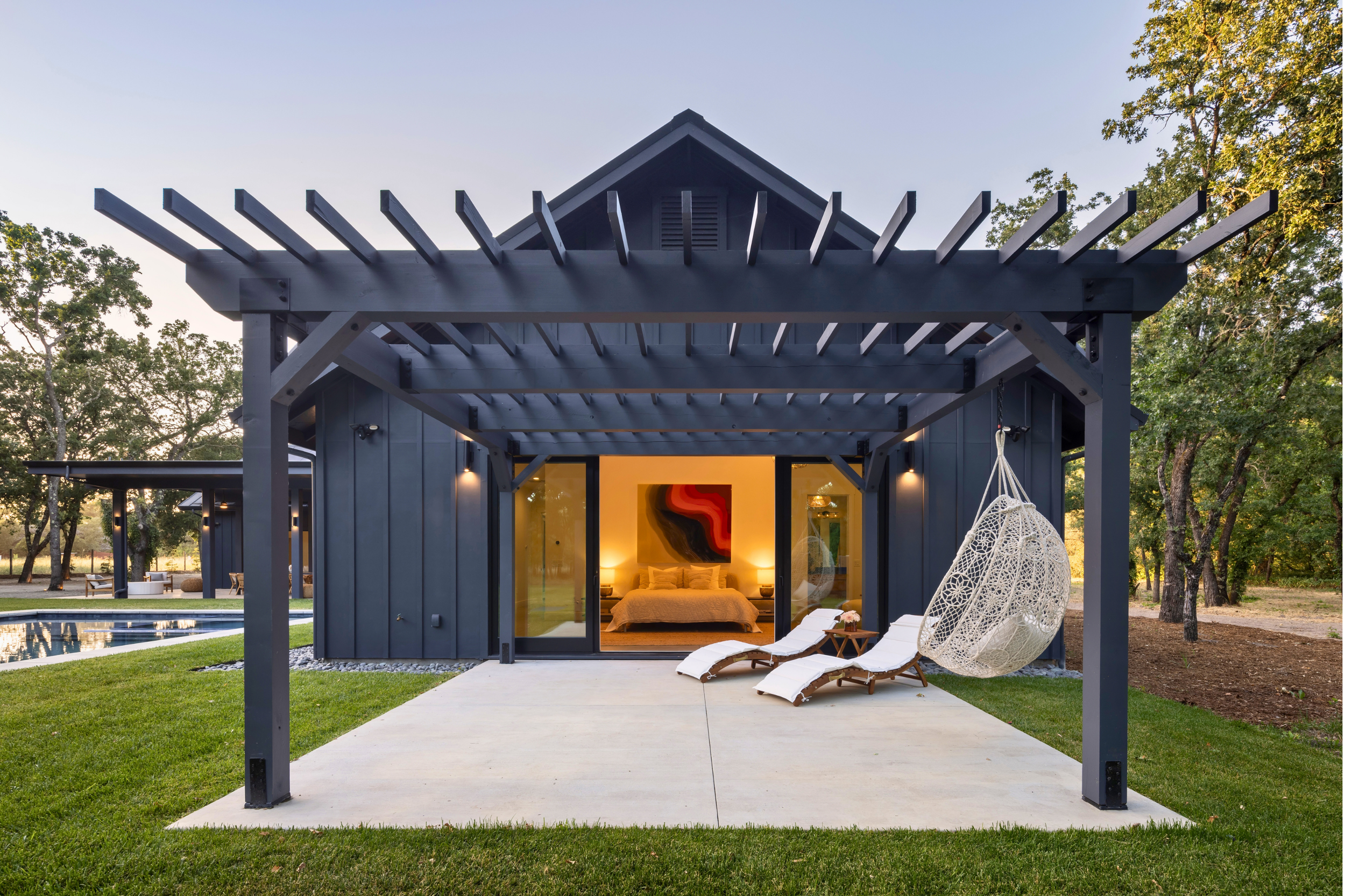 Modern black house with a patio, outdoor hammock chair, lounge chairs, and interior bedroom visible through sliding glass doors, surrounded by green grass and trees.