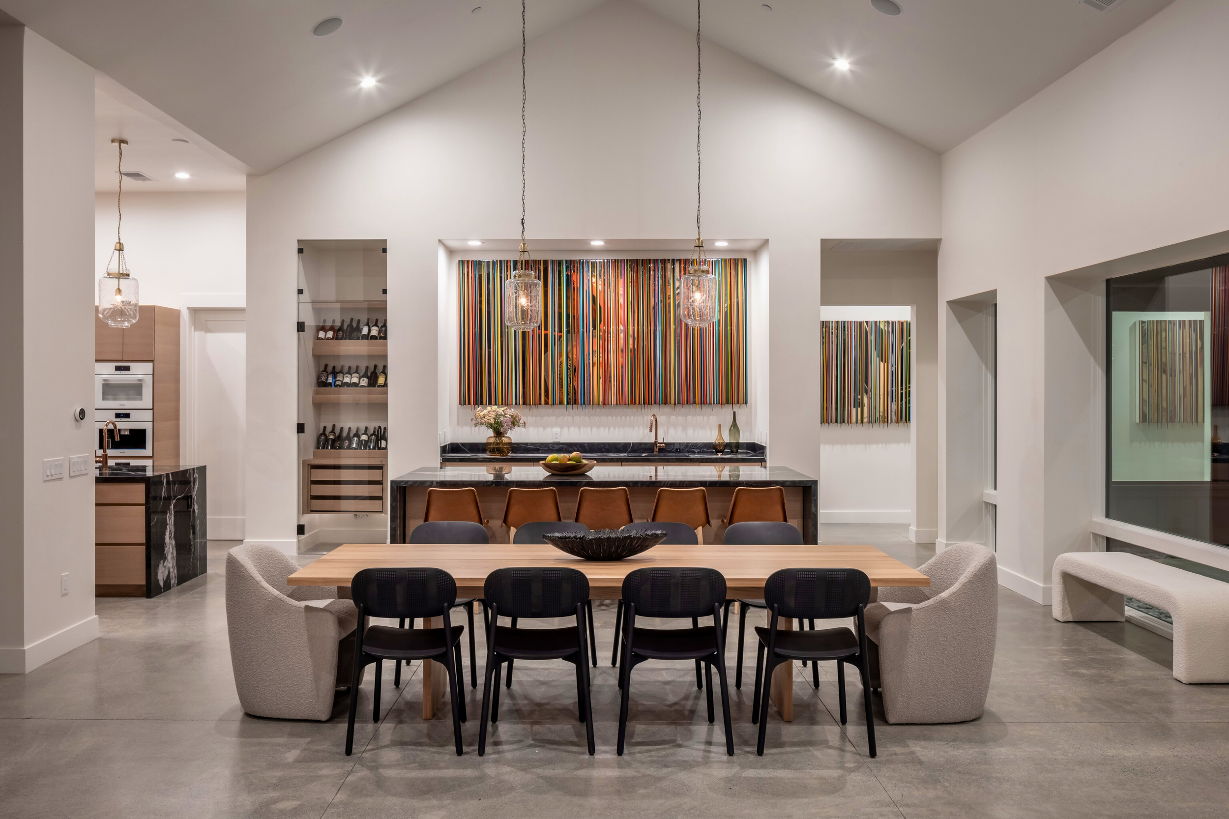 Modern dining area with a large wooden table surrounded by a mix of upholstered and black chairs. Behind the table is a bar counter with colorful abstract art and pendant lights hanging above. The room features white walls, large windows, and polishe
