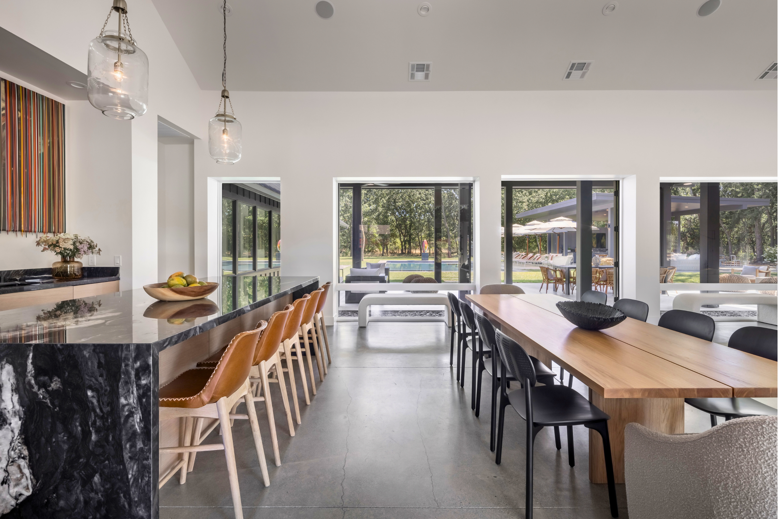 Modern dining area with a black marble countertop bar with brown leather chairs, a wooden dining table with black chairs, and large windows showing an outdoor patio and pool area.
