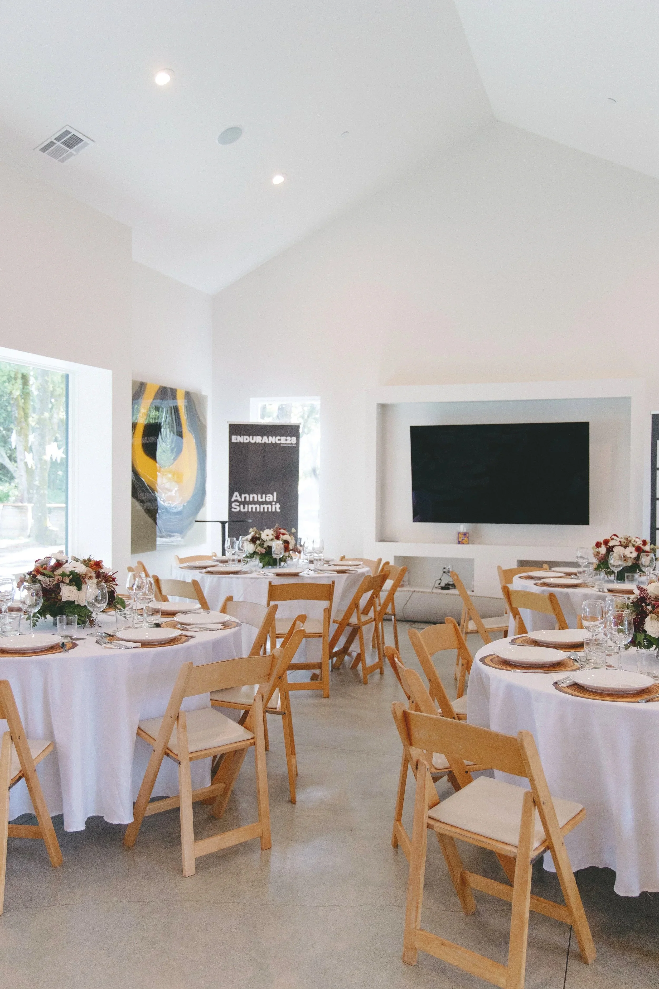 Elegant event room with round tables covered in white tablecloths, decorated with floral centerpieces, and surrounded by wooden chairs, set for a meal during daytime.