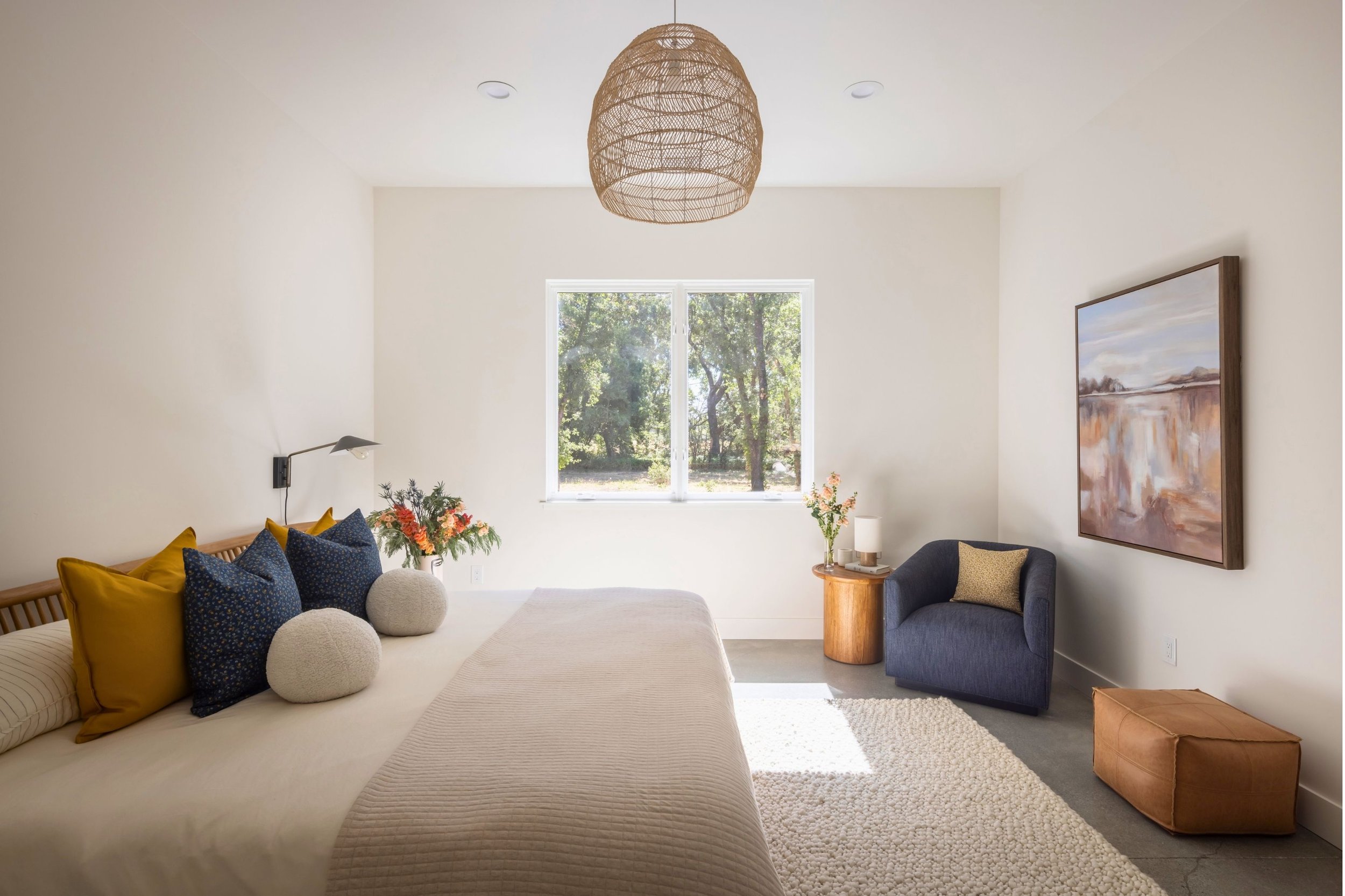 Bright bedroom with a large bed decorated with yellow and navy pillows, a window showing trees outside, a blue armchair with a pillow, a wooden side table with a lamp and flowers, framed artwork on the wall, a ottoman, and a textured white rug.