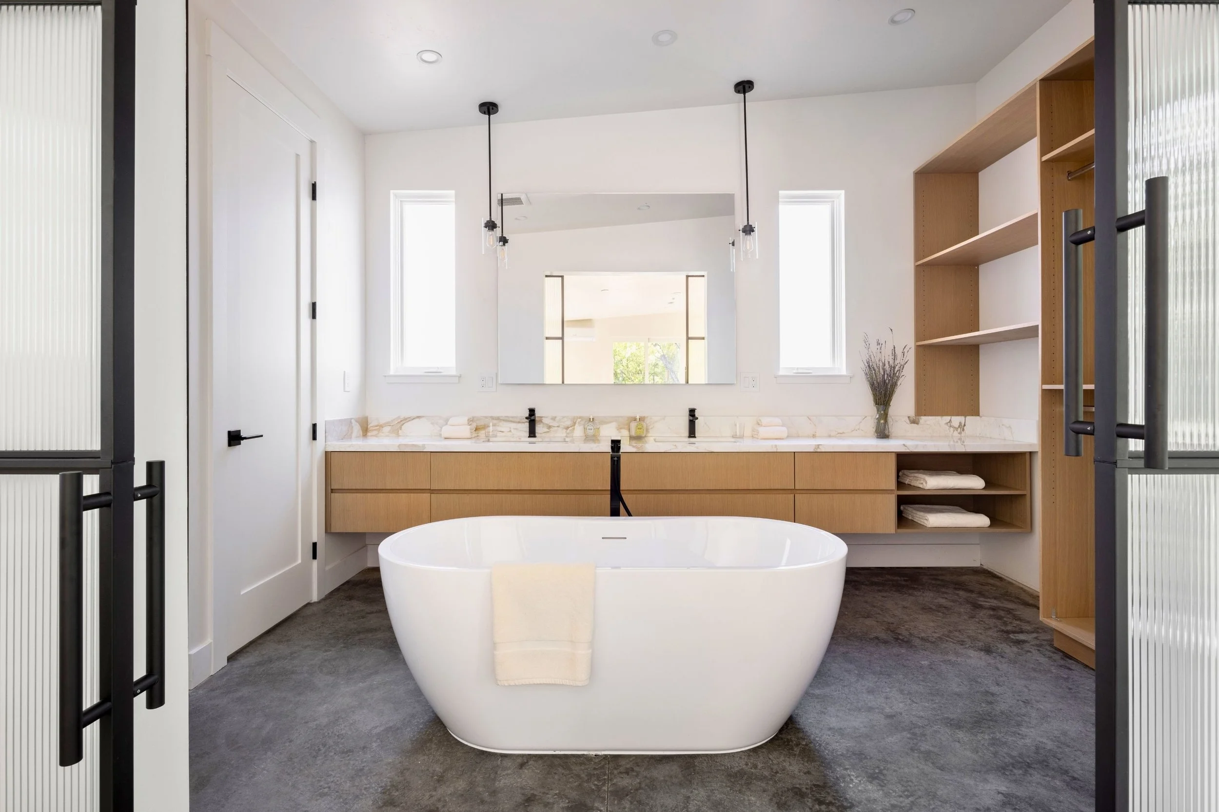 Modern bathroom with a freestanding bathtub, dual sinks with marble countertop, wooden cabinets, large mirror, two small windows, open shelving with towels, and pendant lights.