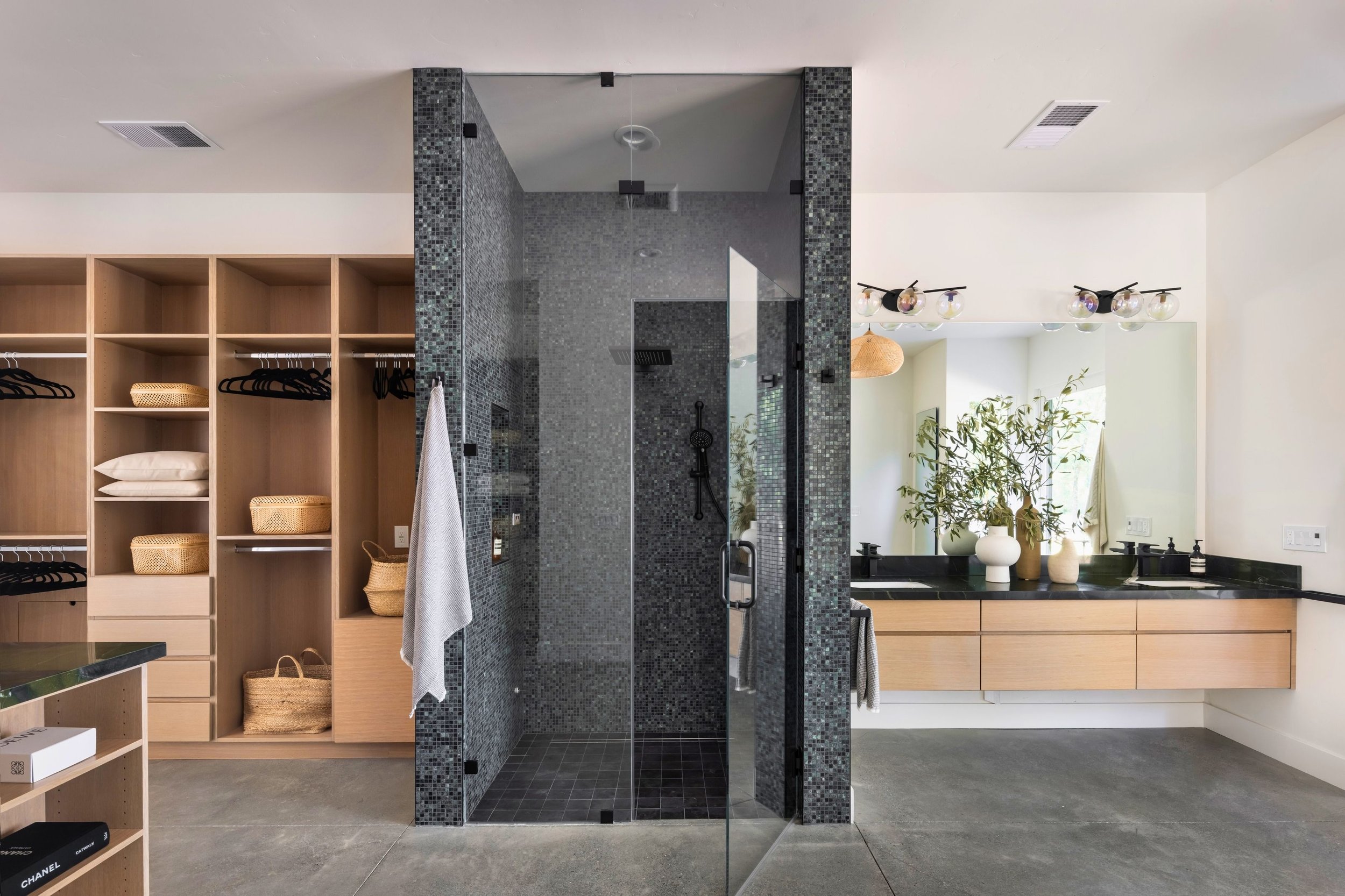 Modern bathroom featuring a glass-enclosed shower with dark mosaic tiles, a floating wooden vanity with a large mirror, and built-in open shelves with towels and baskets. Decor includes potted plants and minimalist lighting fixtures.