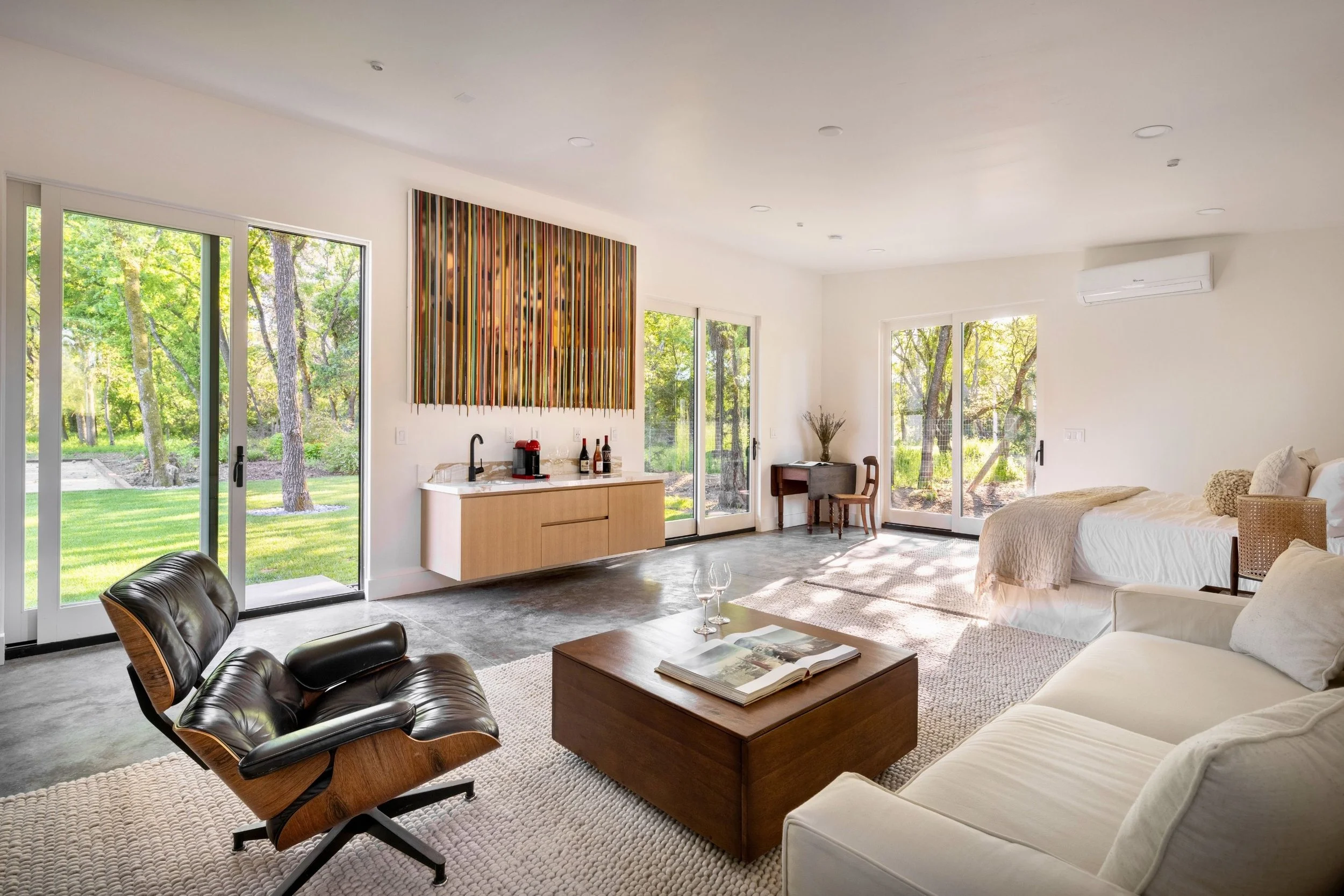 Bright living room with sliding glass doors opening to a green outdoor area, modern furniture including a black leather lounge chair, a wooden coffee table with an open book and two wine glasses, a bed, a small writing desk, colorful abstract wall ar