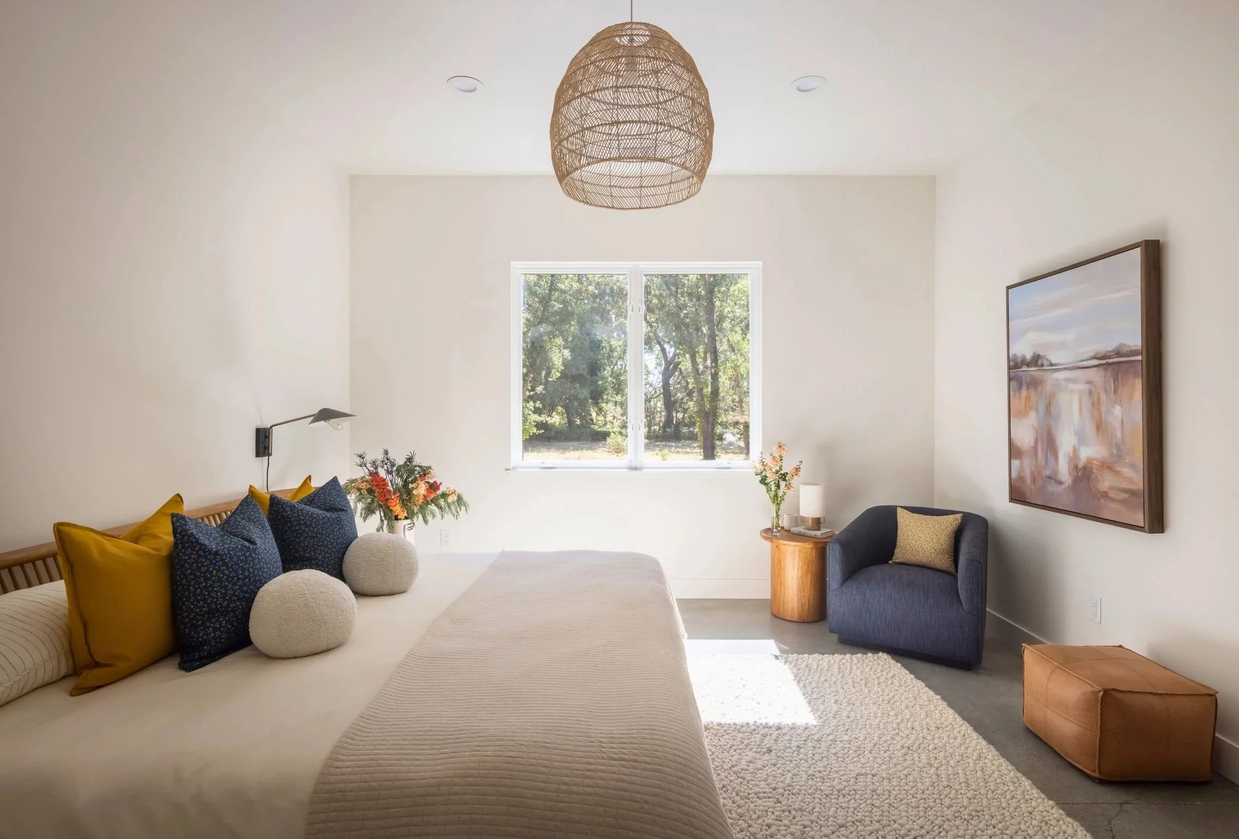 Bright bedroom with neutral walls, a large window showing a green outdoor view, a bed with mustard, navy, and textured pillows, a bedside flowers, a blue armchair with a pillow, a small wooden side table with a lamp, a large framed landscape painting, a brown ottoman, and a cream rug.